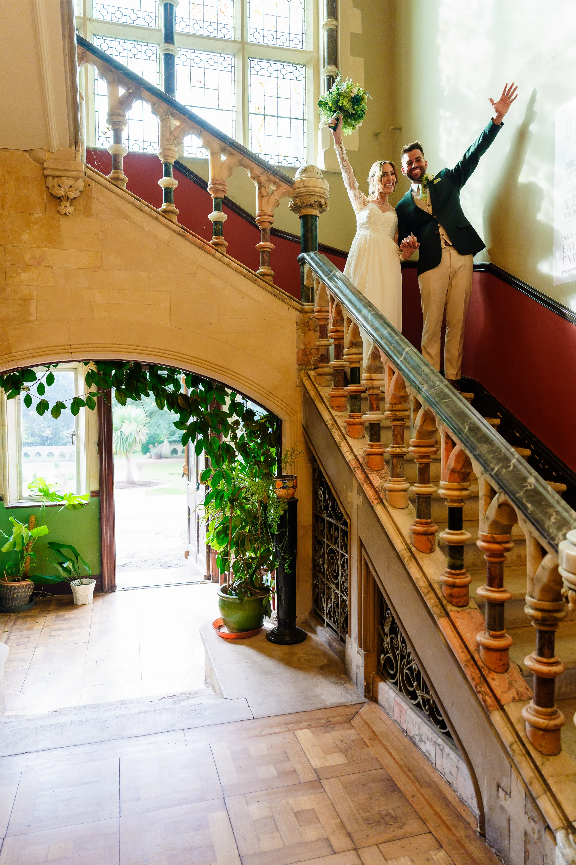 A newlywed couple celebrates by raising their hands on a grand stone staircase inside a historic country house venue.