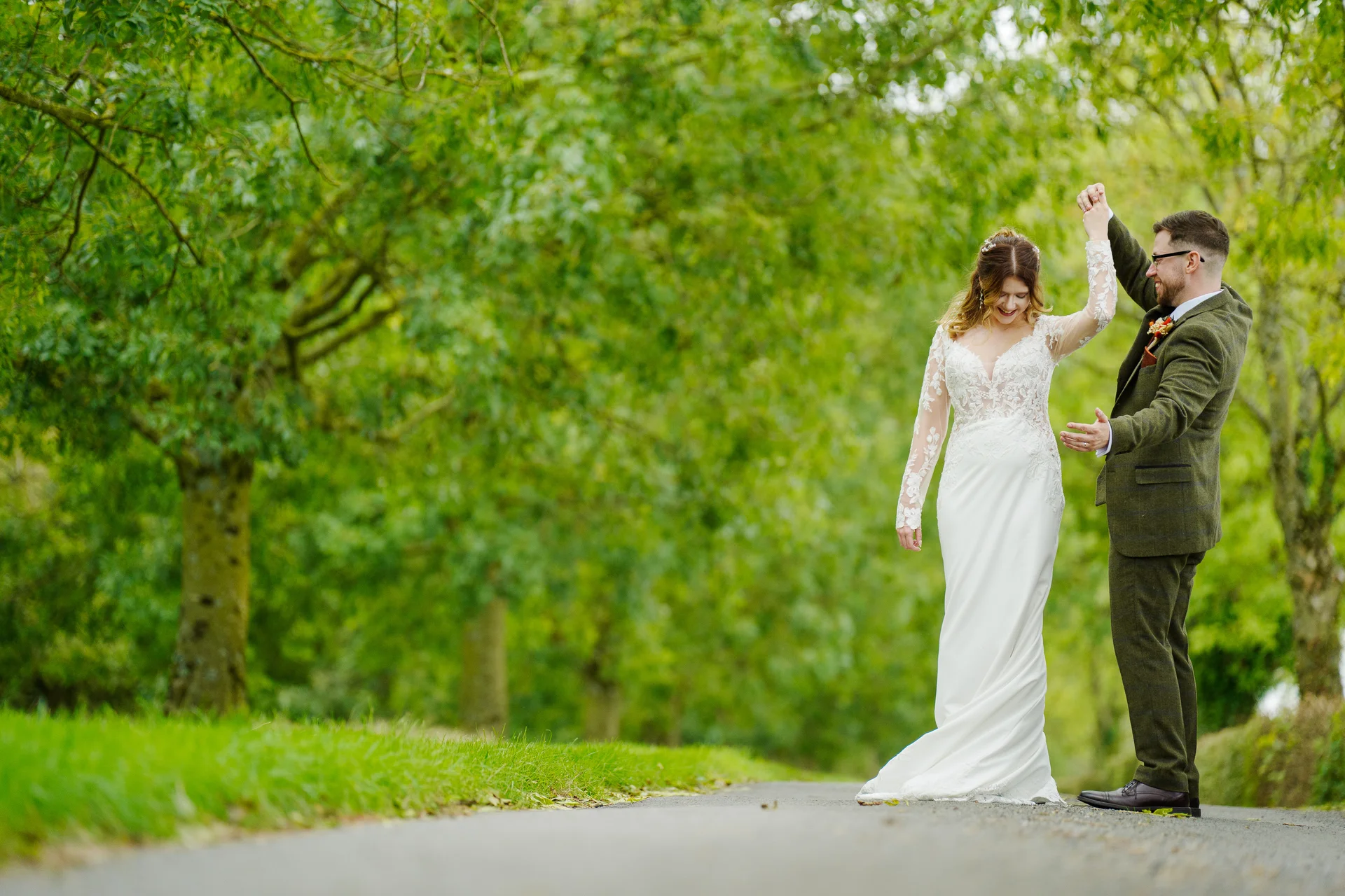 A newlywed couple shares a joyful dance along a country lane lined with lush green trees, with the bride wearing a long-sleeved lace and crepe gown and the groom in a smart tweed suit.