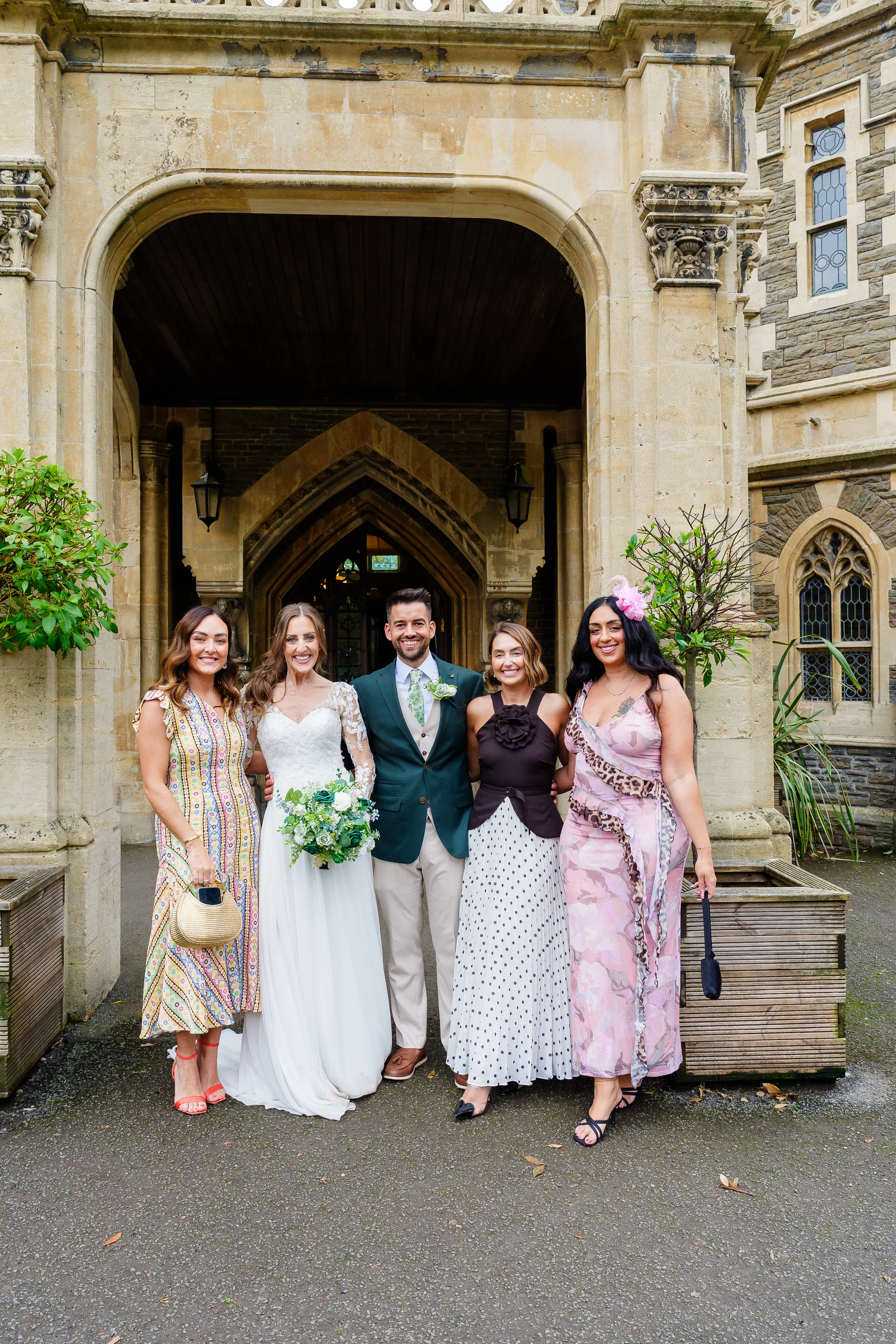 A smiling bride and groom pose outside under a stone archway with four wedding guests, surrounded by historic architecture and lush greenery.