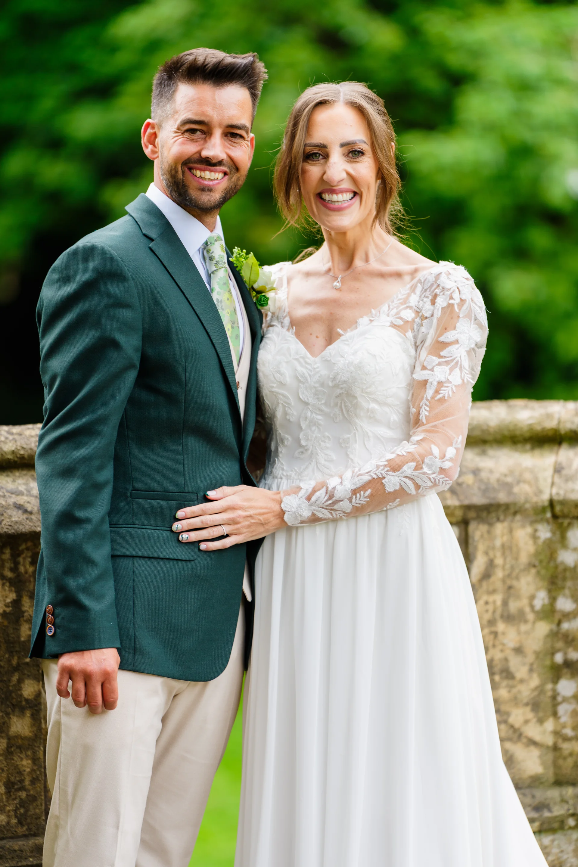 A happy newlywed couple poses together outdoors with the groom wearing a green suit and the bride in a long-sleeve lace wedding dress.