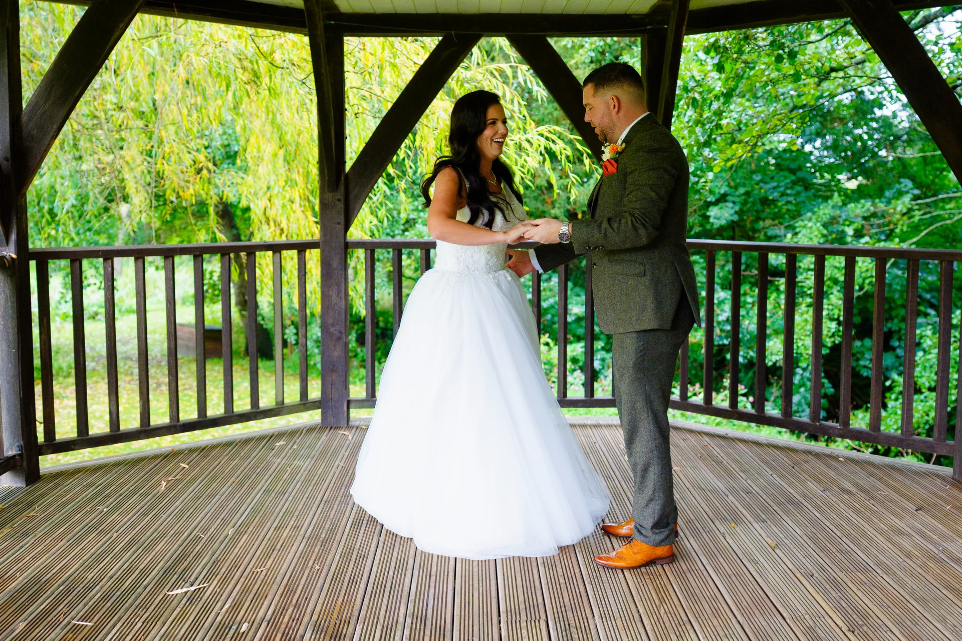 A newlywed couple shares a joyful moment holding hands inside a wooden gazebo surrounded by lush green foliage.