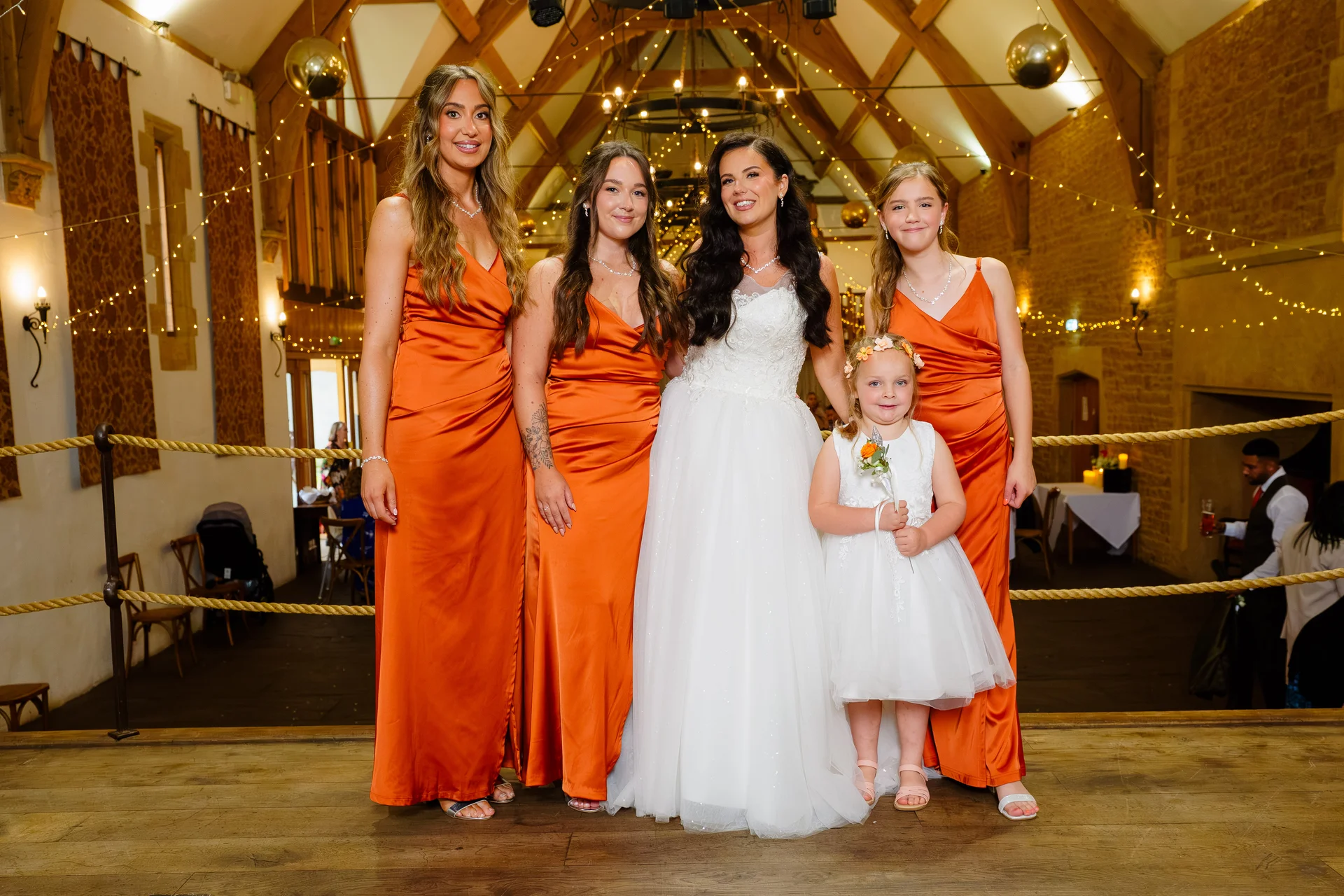 A smiling bride in a white tulle wedding dress poses alongside four bridesmaids wearing vibrant orange satin gowns and a young flower girl inside a rustic barn decorated with fairy lights.