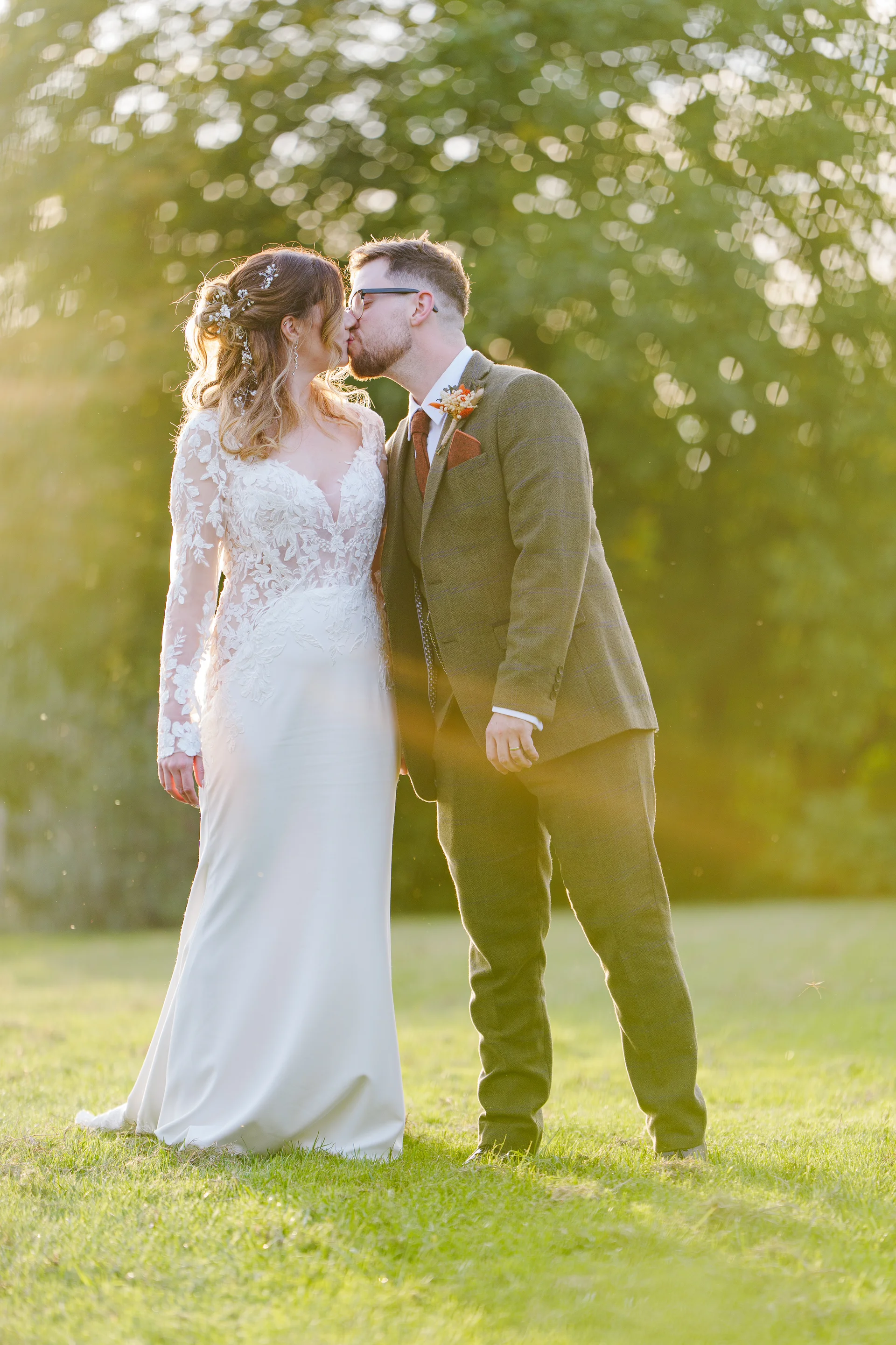 Newlyweds share a romantic kiss outdoors during golden hour, surrounded by a lush green lawn and sunlit foliage.