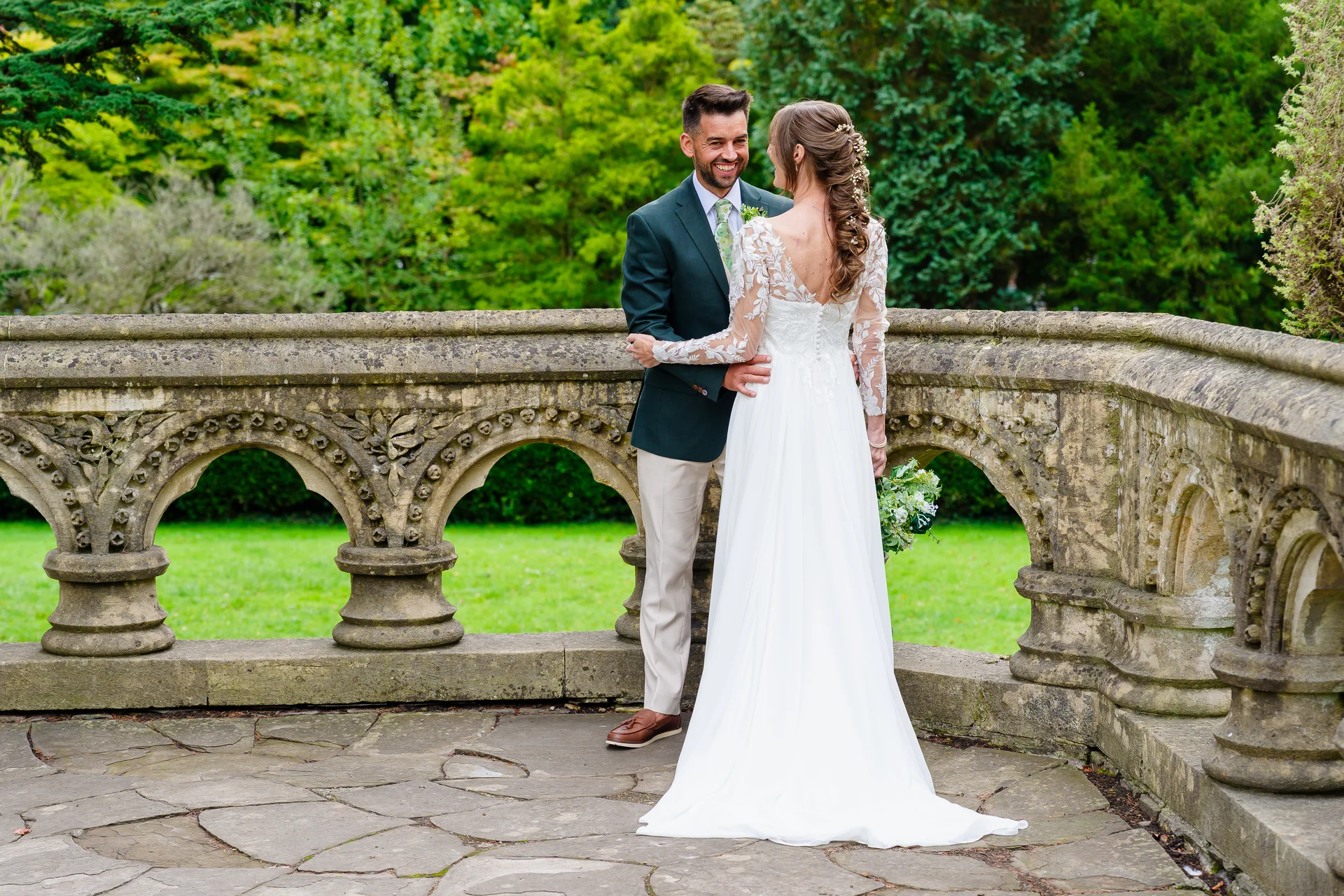 A newlywed couple shares a tender moment on a stone terrace overlooking lush green gardens, with the bride in a long-sleeved lace gown and the groom in a dark green blazer.