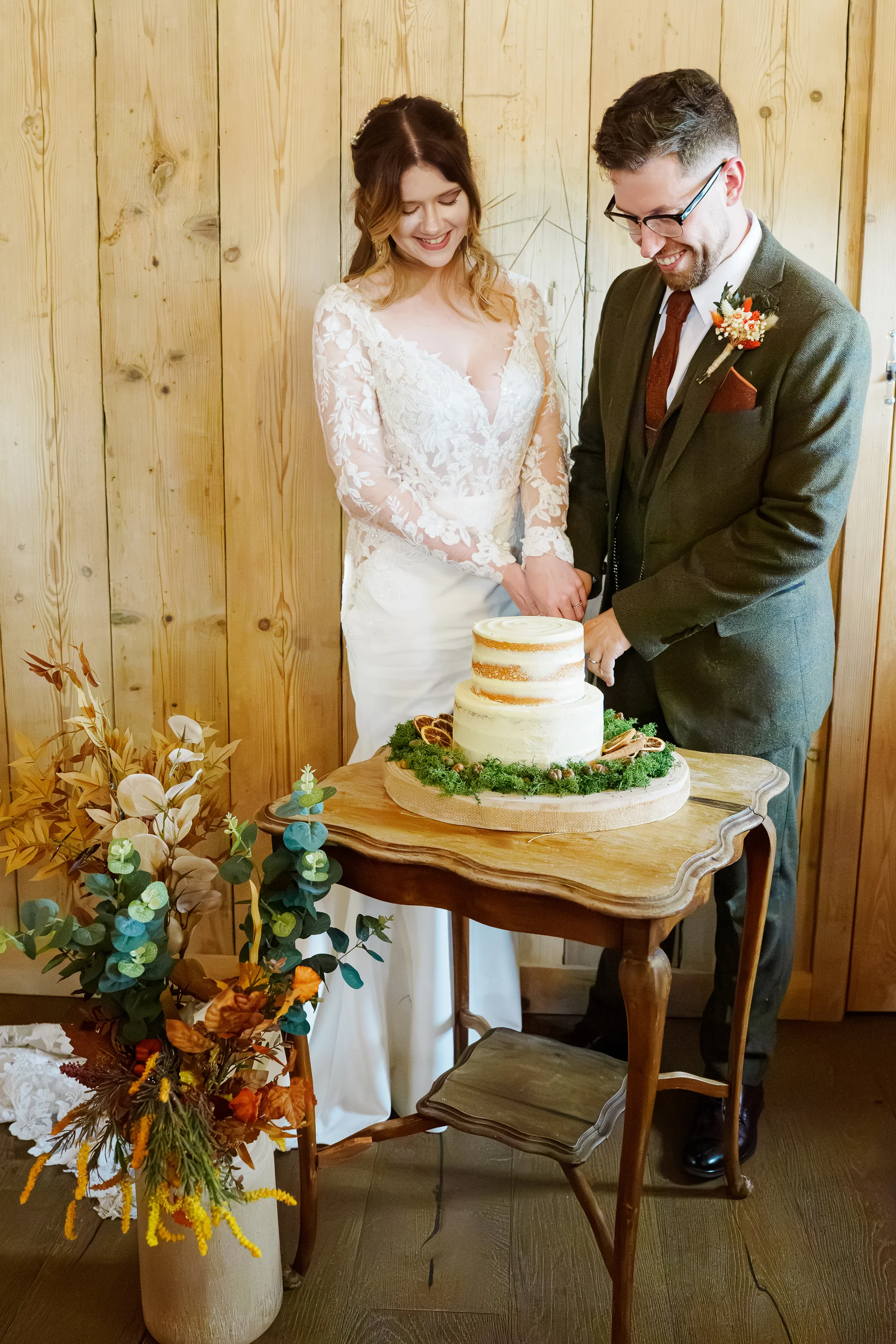 A smiling bride in a long-sleeved lace gown and a groom in a green tweed suit cut a rustic semi-naked wedding cake adorned with moss and dried citrus slices inside a wooden barn.
