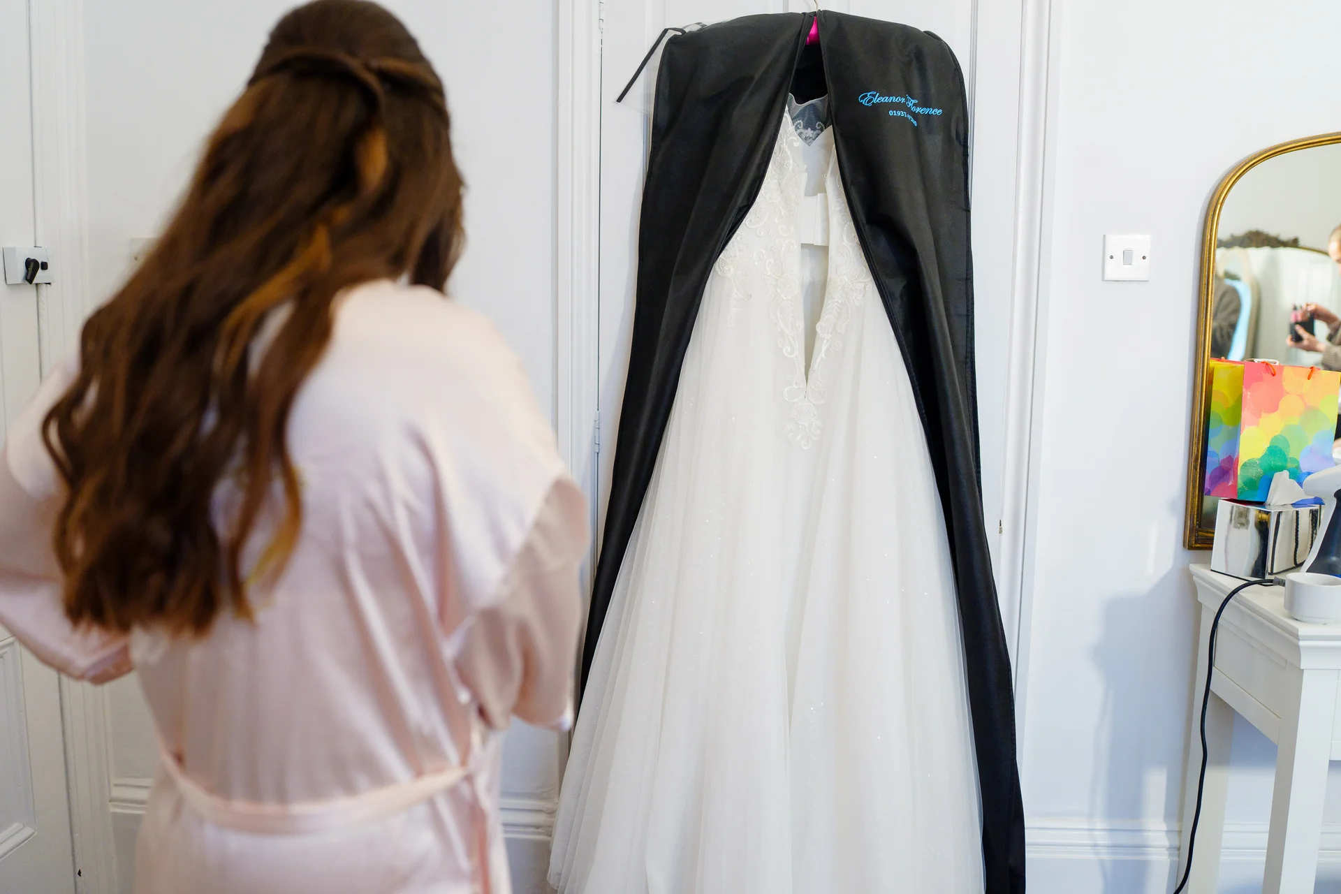 A bride with long brown hair wearing a pink getting ready robe looks at her wedding dress hanging in a black garment bag.