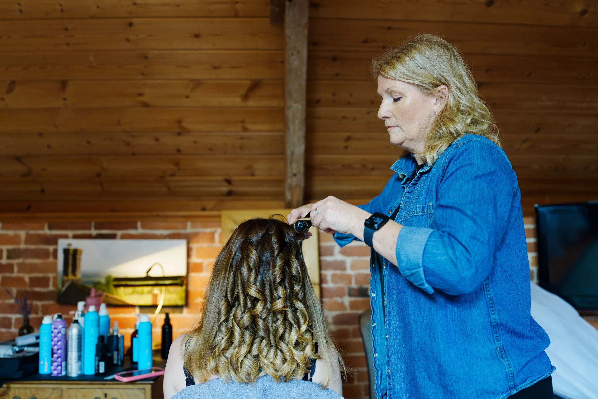 A professional hairstylist carefully styles a bride's long wavy hair inside a rustic barn with exposed brick and wooden panel walls during morning preparations.