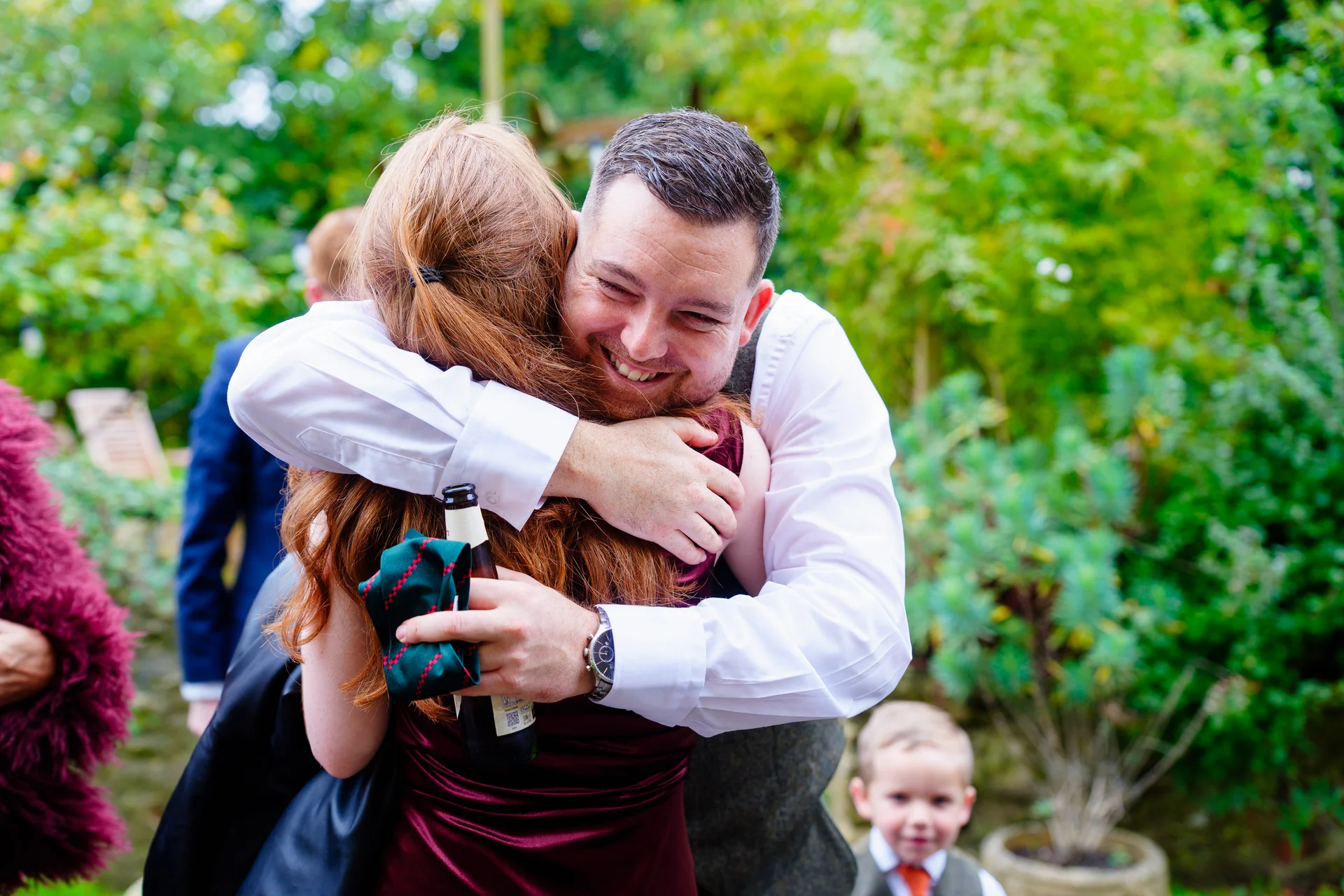 A smiling man in a tweed waistcoat and white shirt warmly embraces a guest outdoors amidst lush greenery during a wedding celebration.