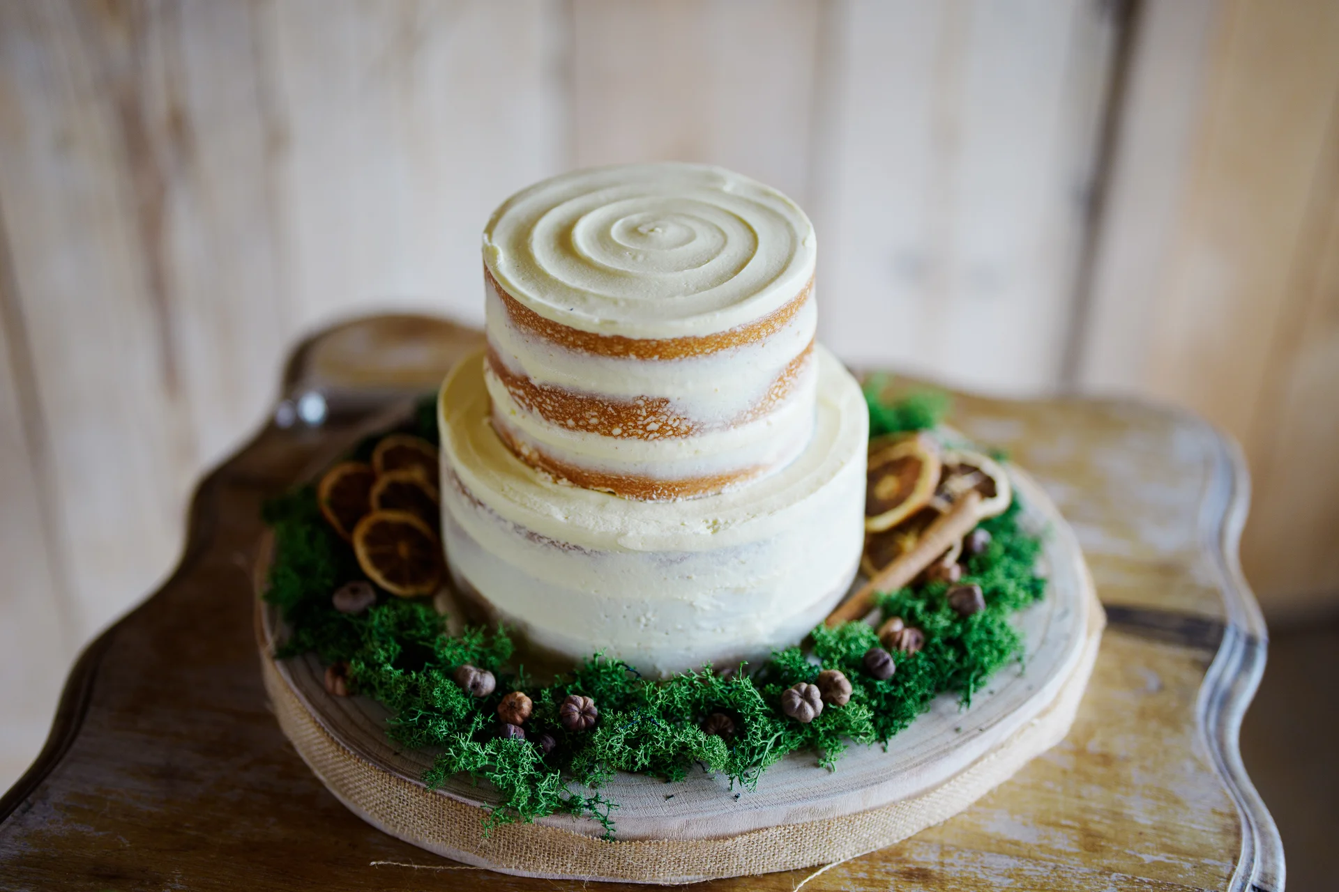 A two-tier semi-naked wedding cake styled on a rustic wood slice surrounded by green moss and dried citrus slices.