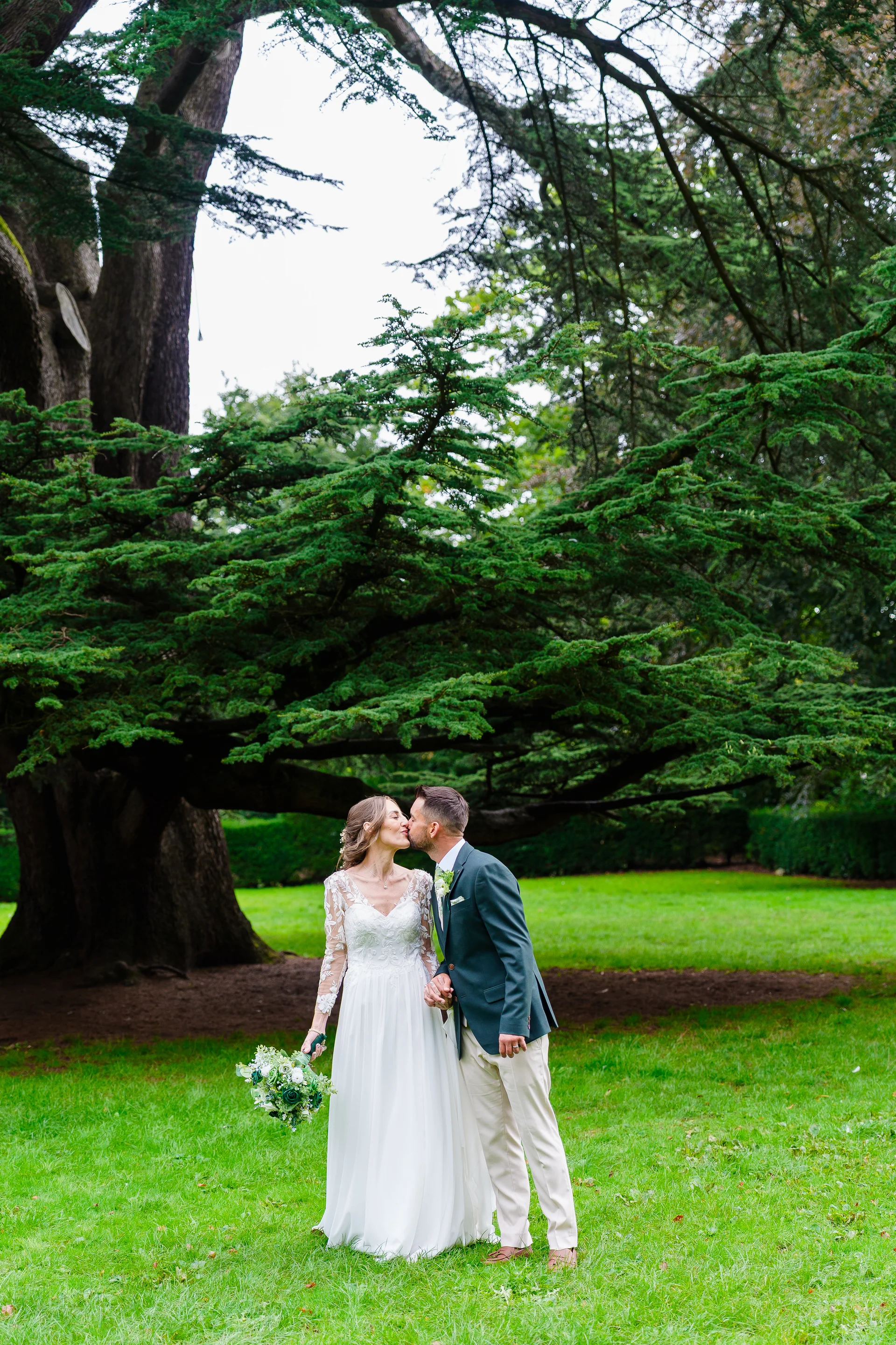 Newlyweds share a tender kiss beneath the sprawling branches of an ancient tree on a lush green lawn.