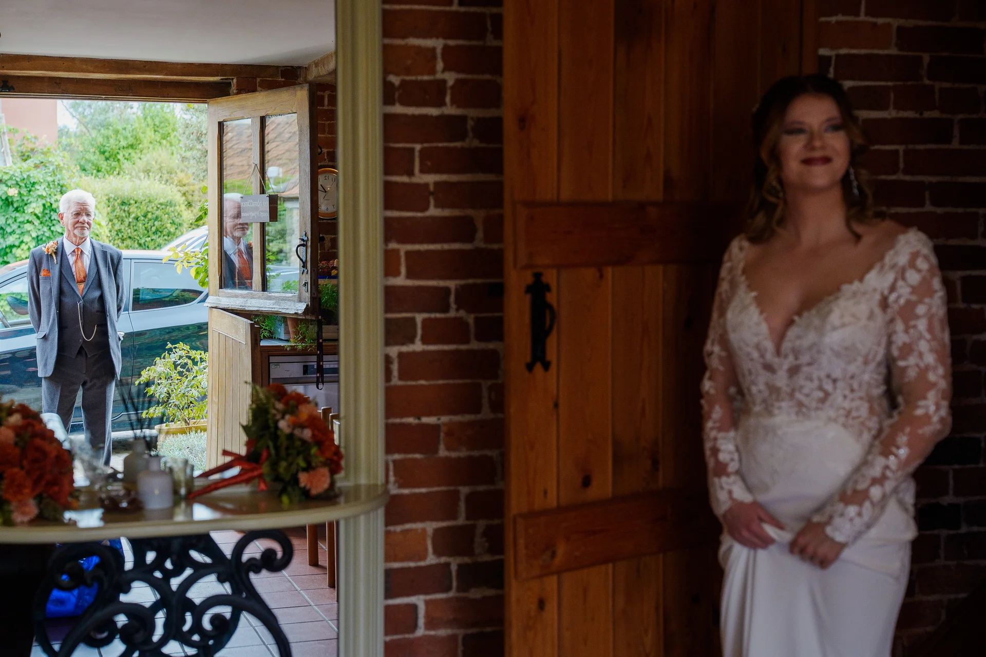 A smiling bride in a lace gown looks towards her father, who stands outside a rustic brick doorway waiting to see her.
