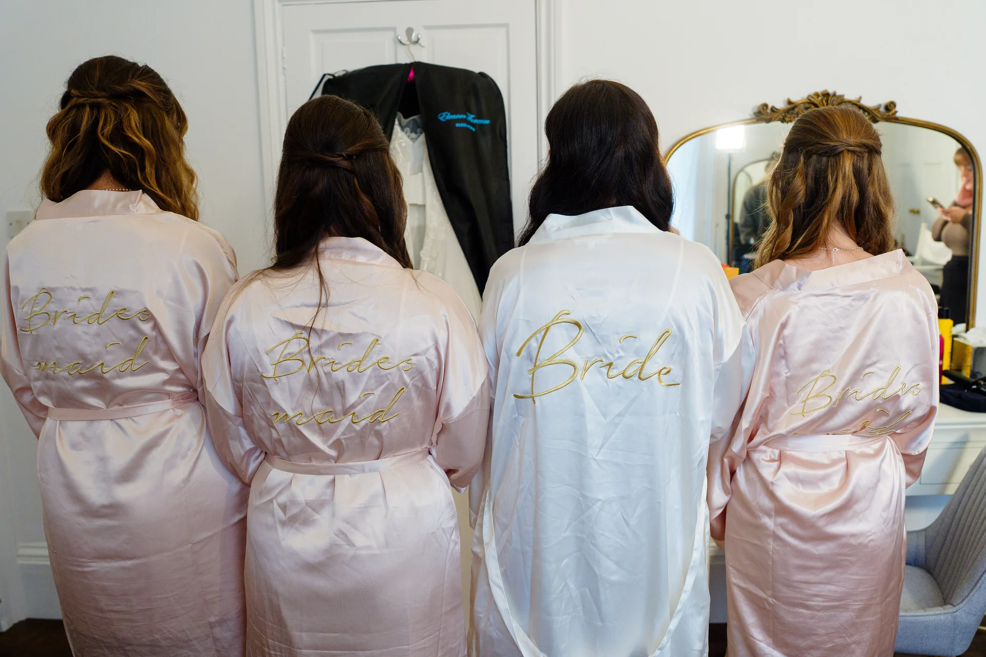 A bride in a white robe and three bridesmaids in matching pink robes stand facing away from the camera during morning preparations.