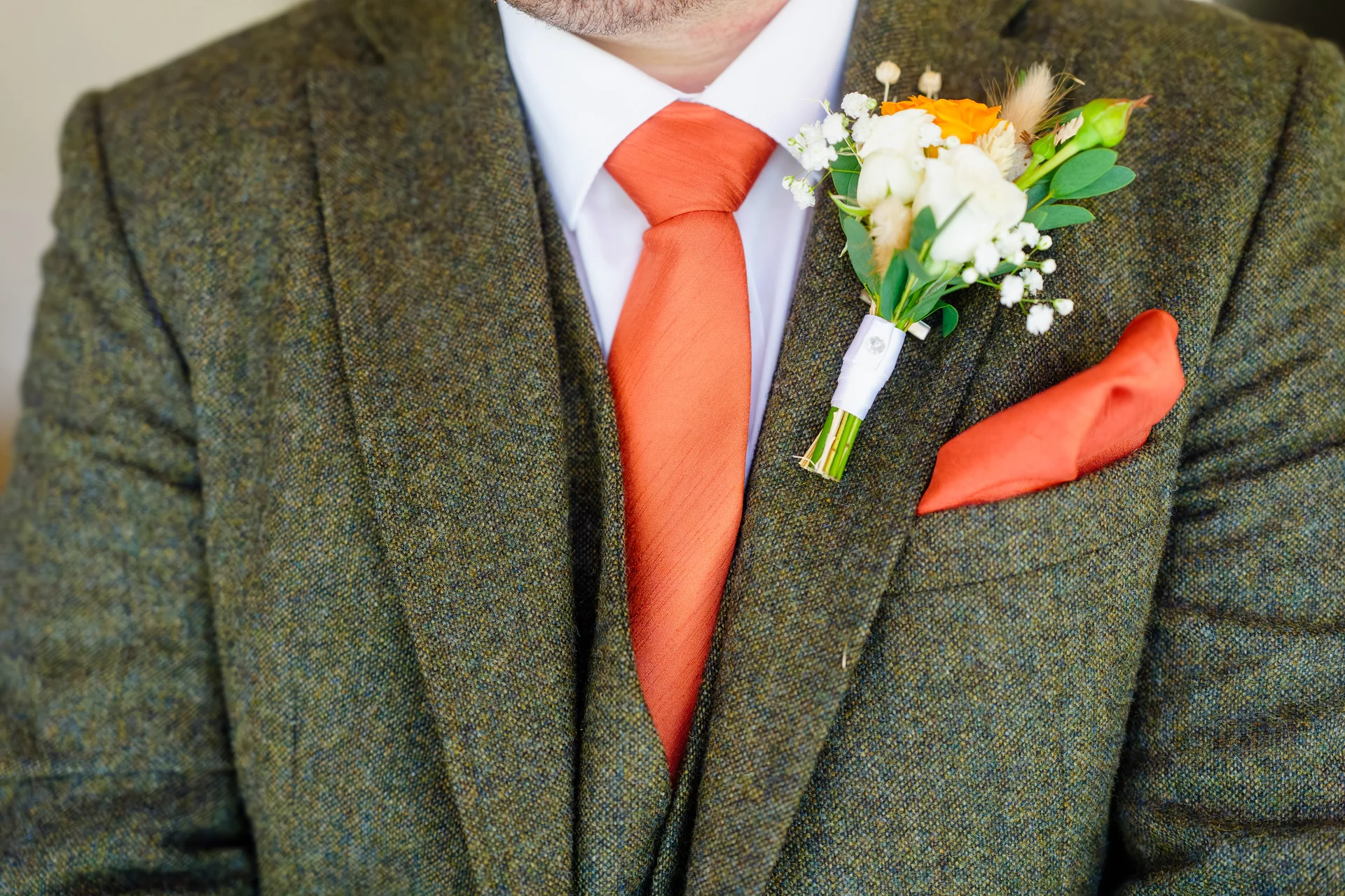 Close-up of a groom wearing a textured tweed three-piece suit paired with a vibrant burnt orange tie, matching pocket square, and coordinating floral buttonhole.