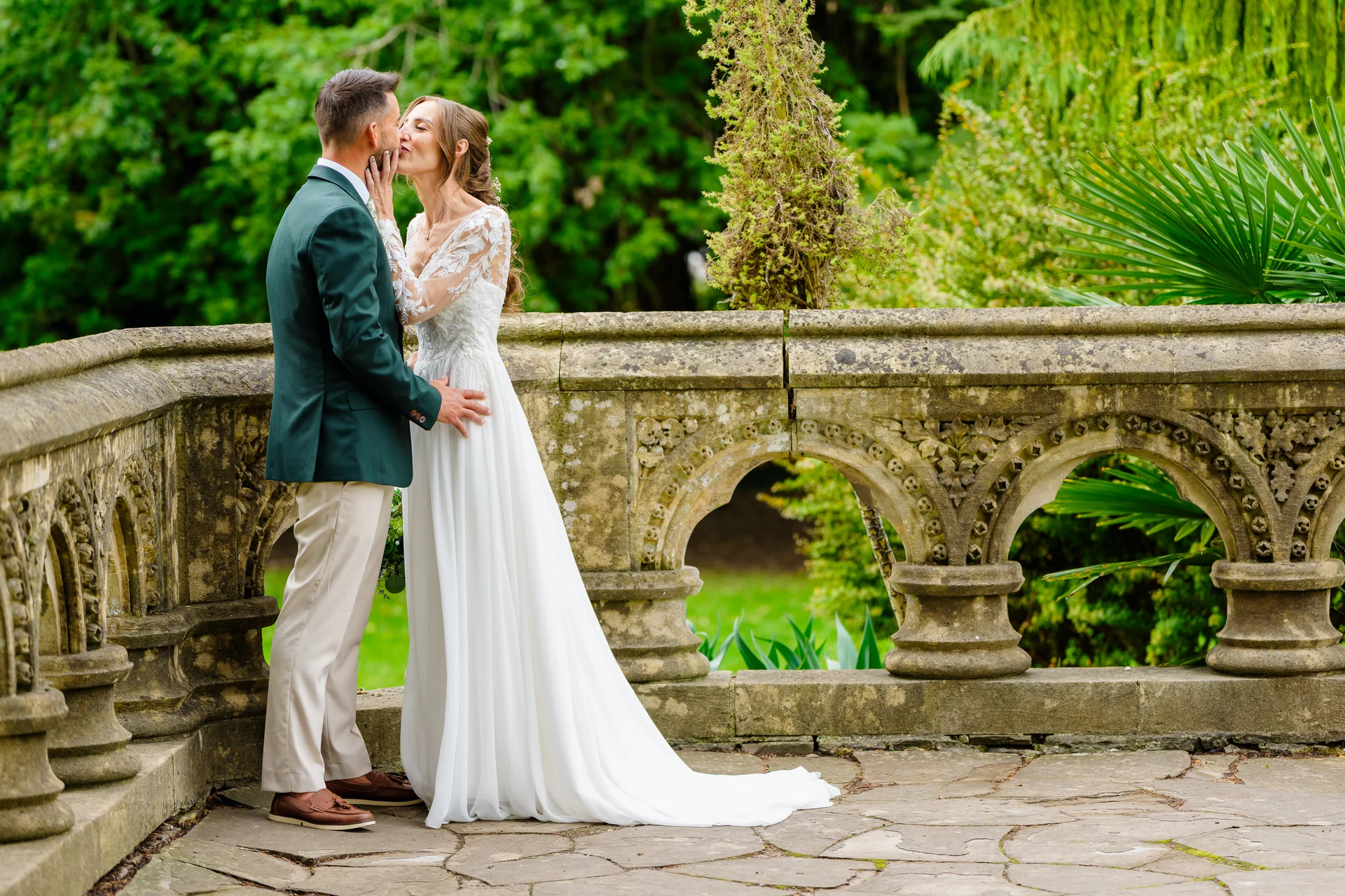 A newlywed couple shares a tender kiss on a stone terrace overlooking lush green gardens during their summer wedding.