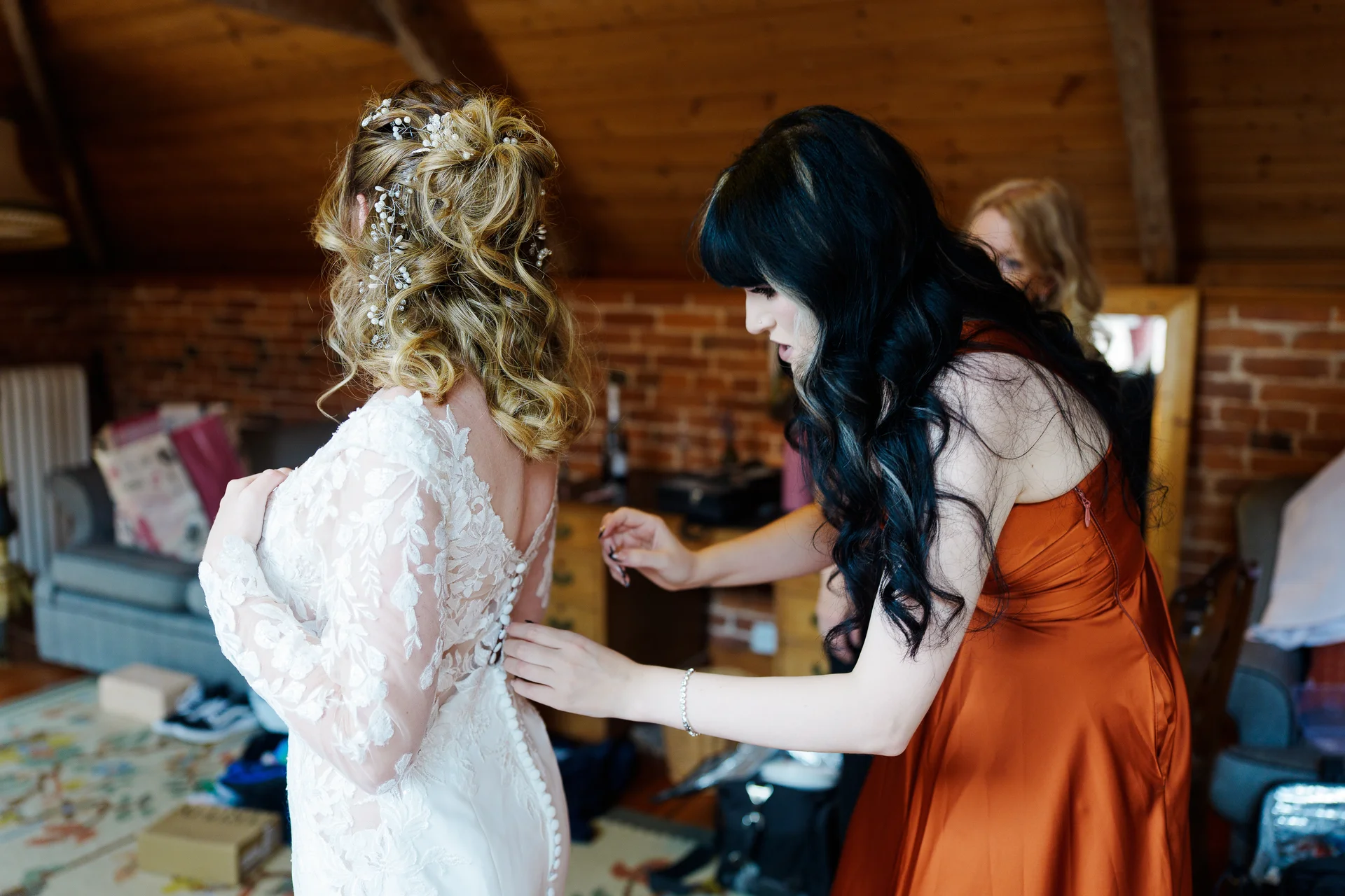 A bridesmaid carefully fastens the button-up back of a bride's intricate lace long-sleeve wedding dress in a rustic room with exposed brick walls.