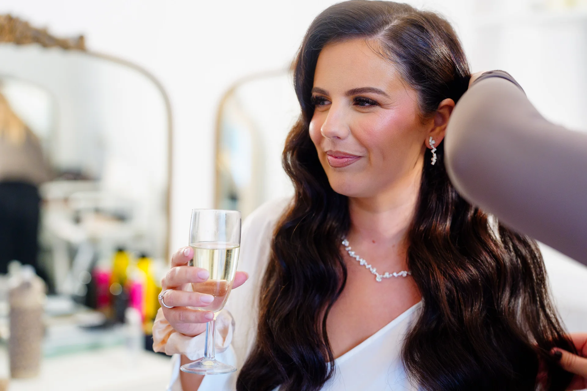 A smiling brunette bride enjoys a glass of champagne while having her long hair styled during bridal preparations.