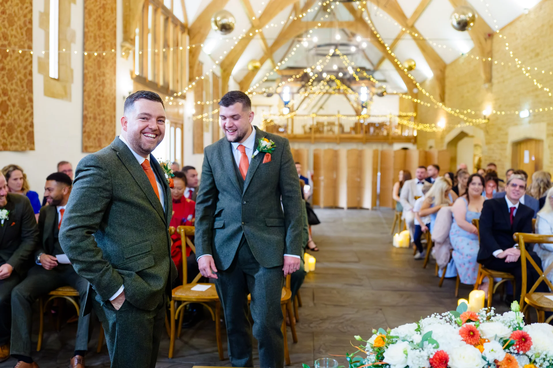 Two grooms in matching green tweed suits smile joyfully at the altar inside a rustic oak-framed barn adorned with fairy lights and floral arrangements.