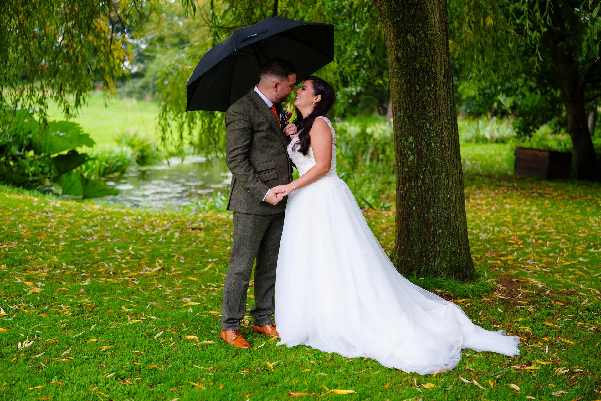 A newlywed couple shares a tender nose-to-nose moment under a black umbrella while standing on a lush green lawn near a wooded pond.
