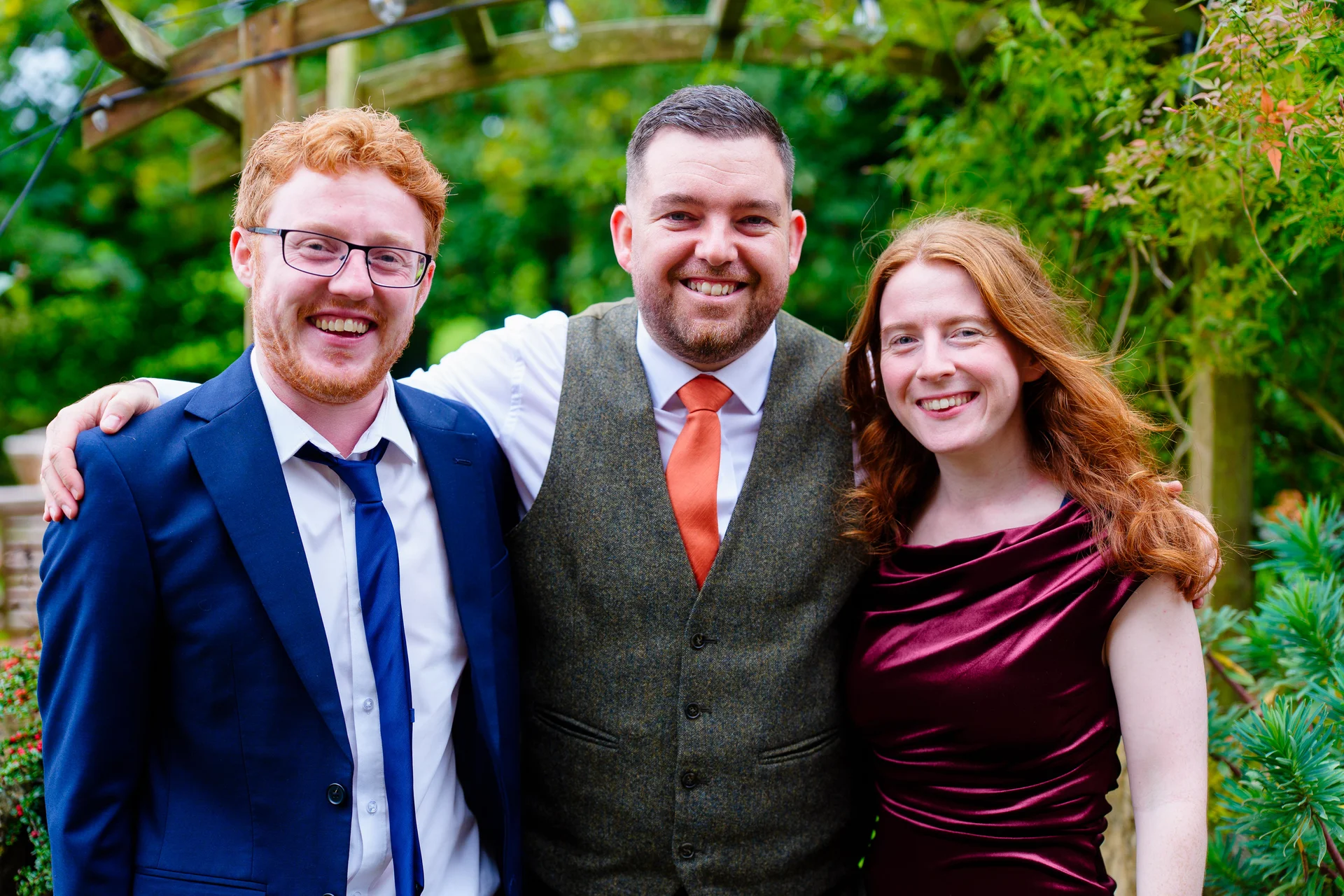Three smiling wedding guests pose together outdoors with greenery in the background, featuring a man in a navy suit, a man in a tweed waistcoat with an orange tie, and a woman in a velvet burgundy dress.