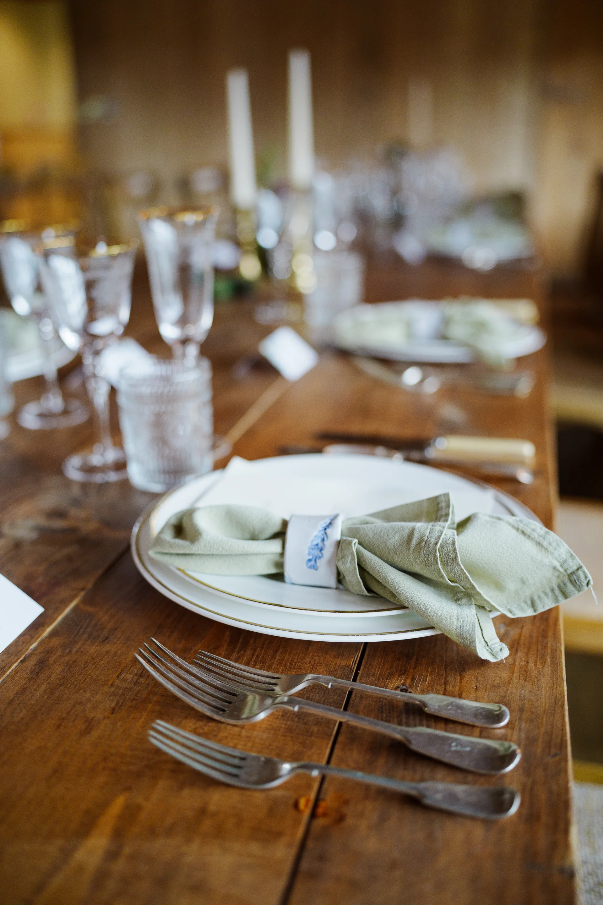 An elegantly styled wedding banquet table features sage green linen napkins held by patterned napkin rings, layered white-and-gold plates, and polished silverware resting on a dark wooden surface.