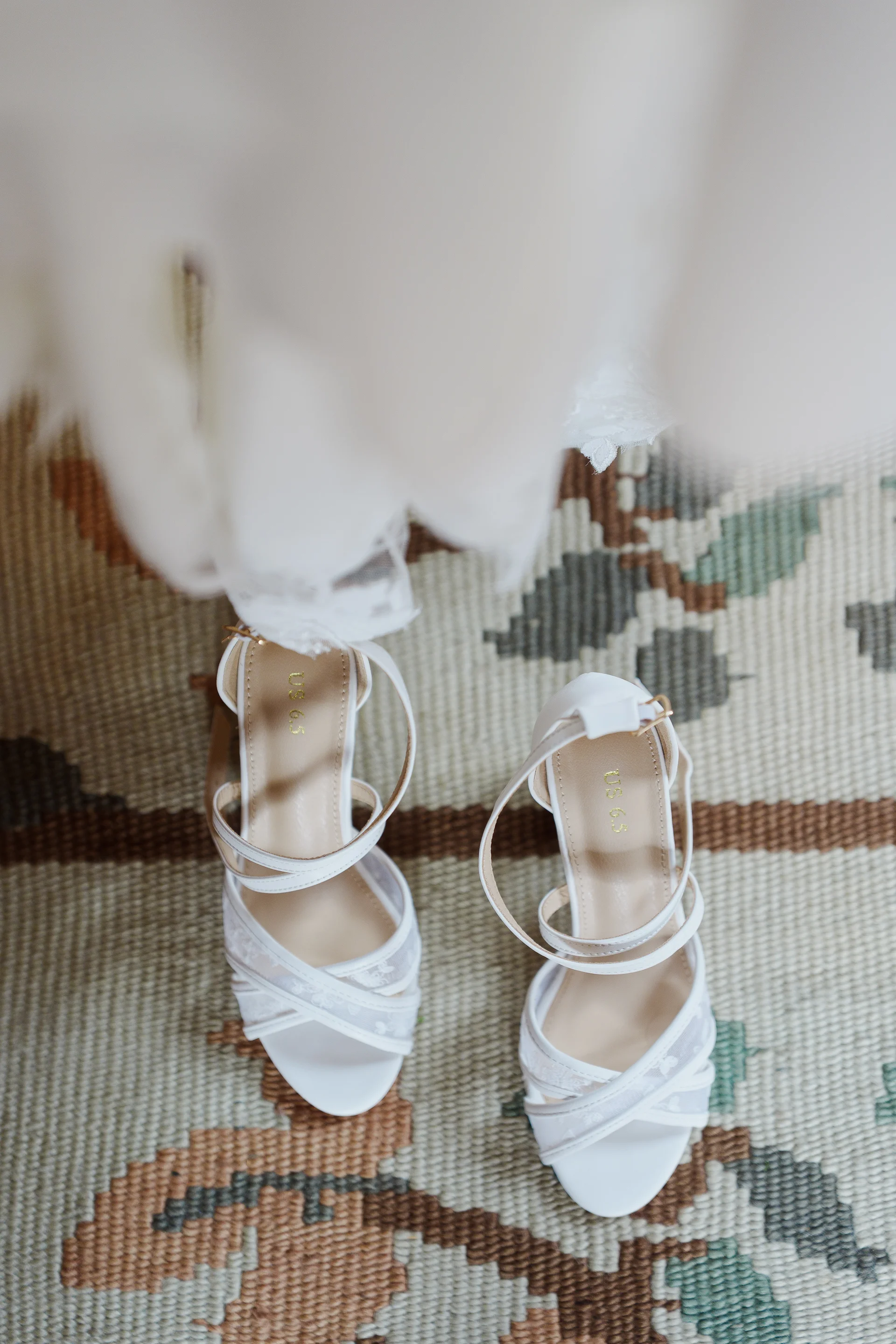 A high-angle shot captures a pair of elegant white bridal strap heels resting on a patterned rug beneath a hanging wedding dress.