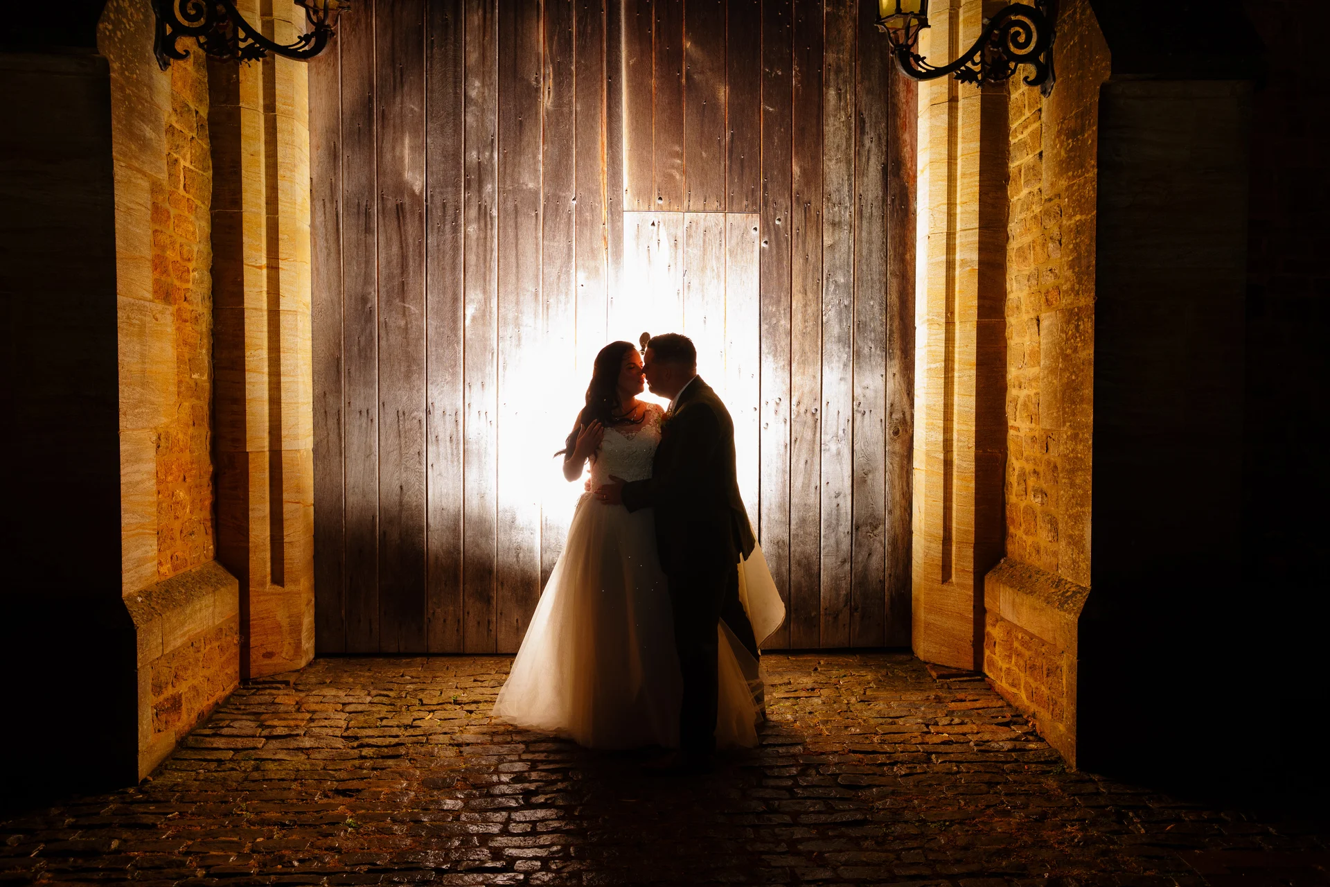 A newlywed couple shares a tender kiss under dramatic backlighting in front of massive wooden doors within a historic stone archway at night.