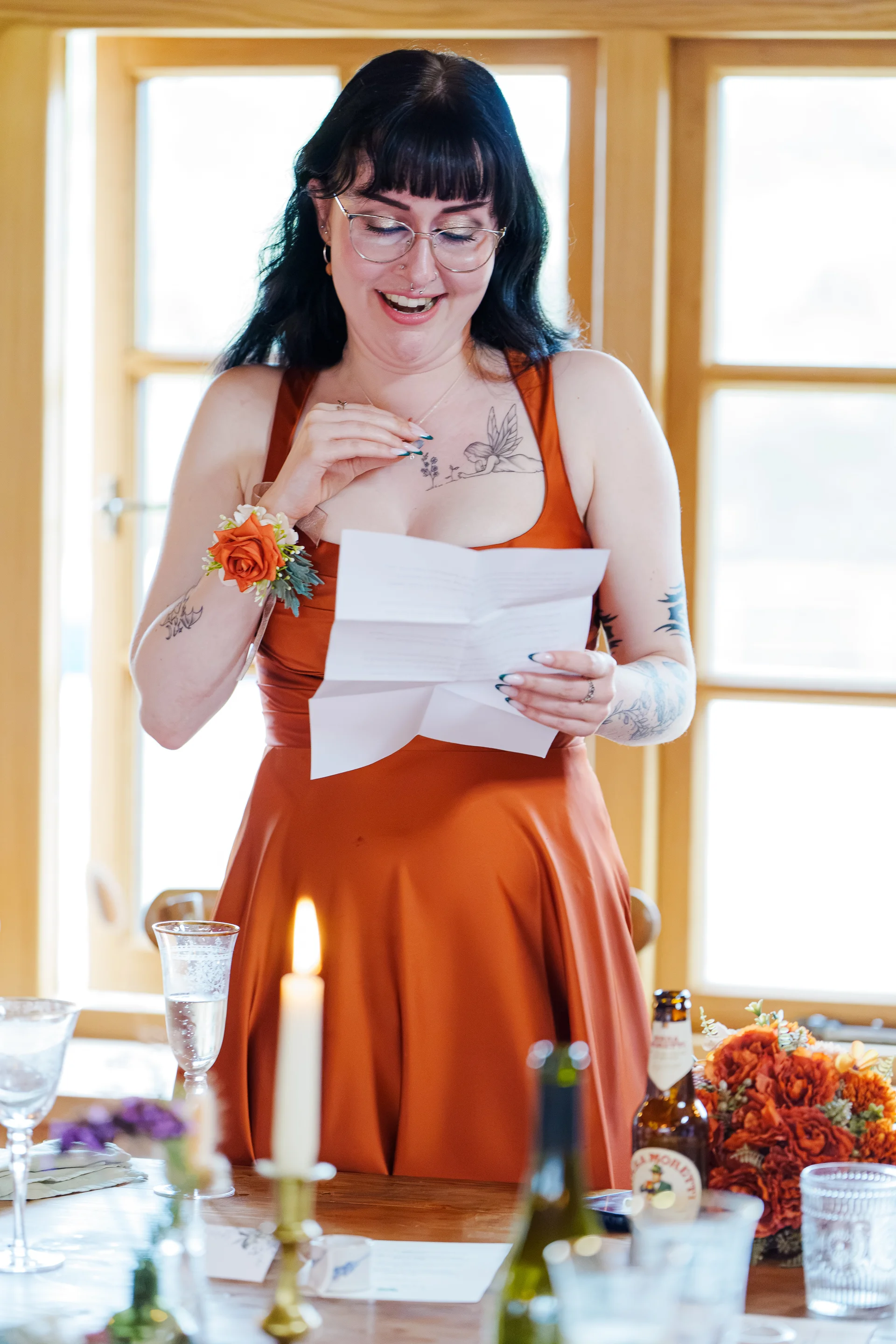 A smiling bridesmaid with dark hair and fringe reads a letter at a wooden table decorated with candles and orange florals.