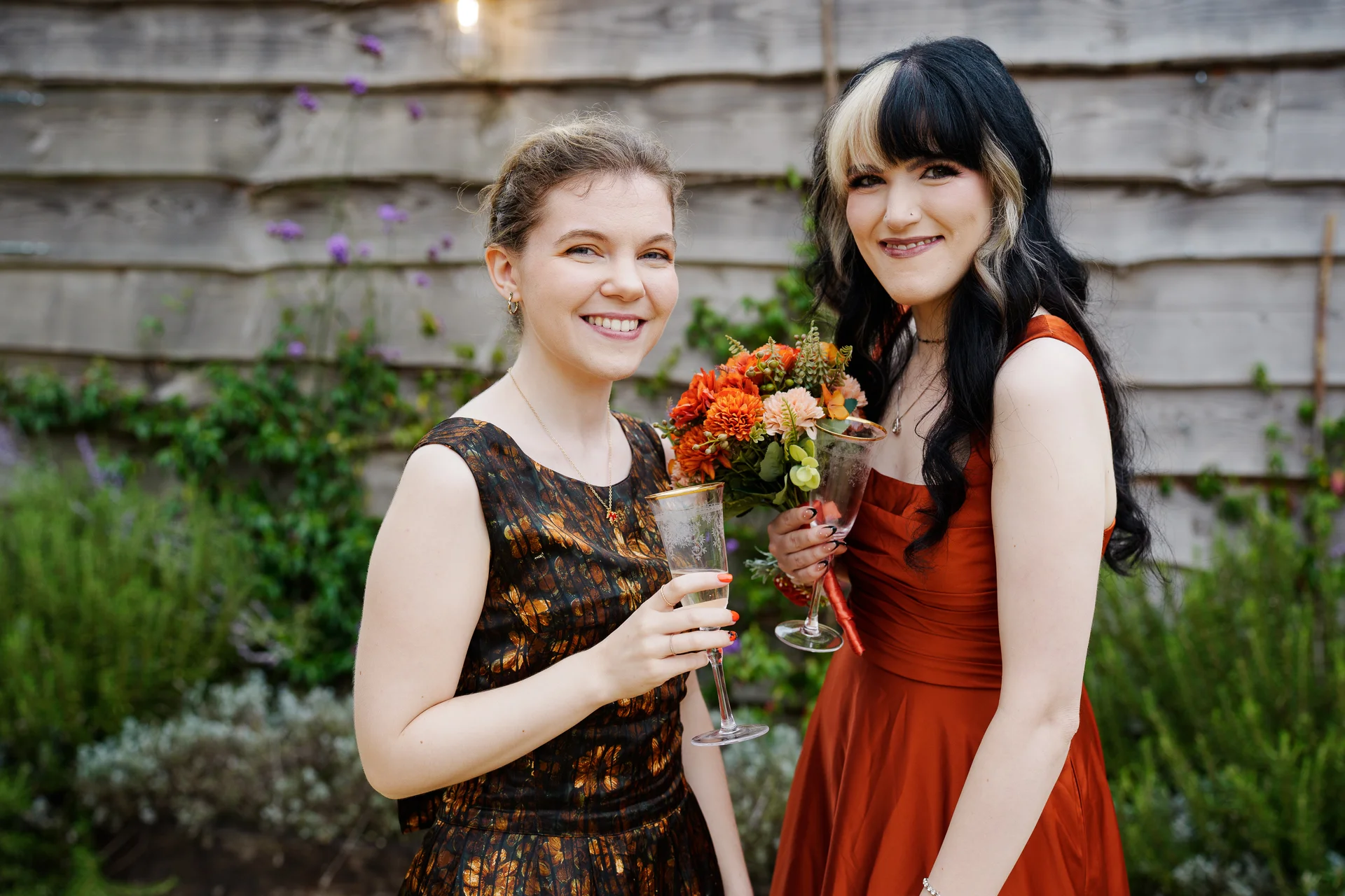 Two smiling brides stand outside against a wooden wall, holding champagne glasses and a vibrant autumn-toned floral bouquet during their wedding celebration.