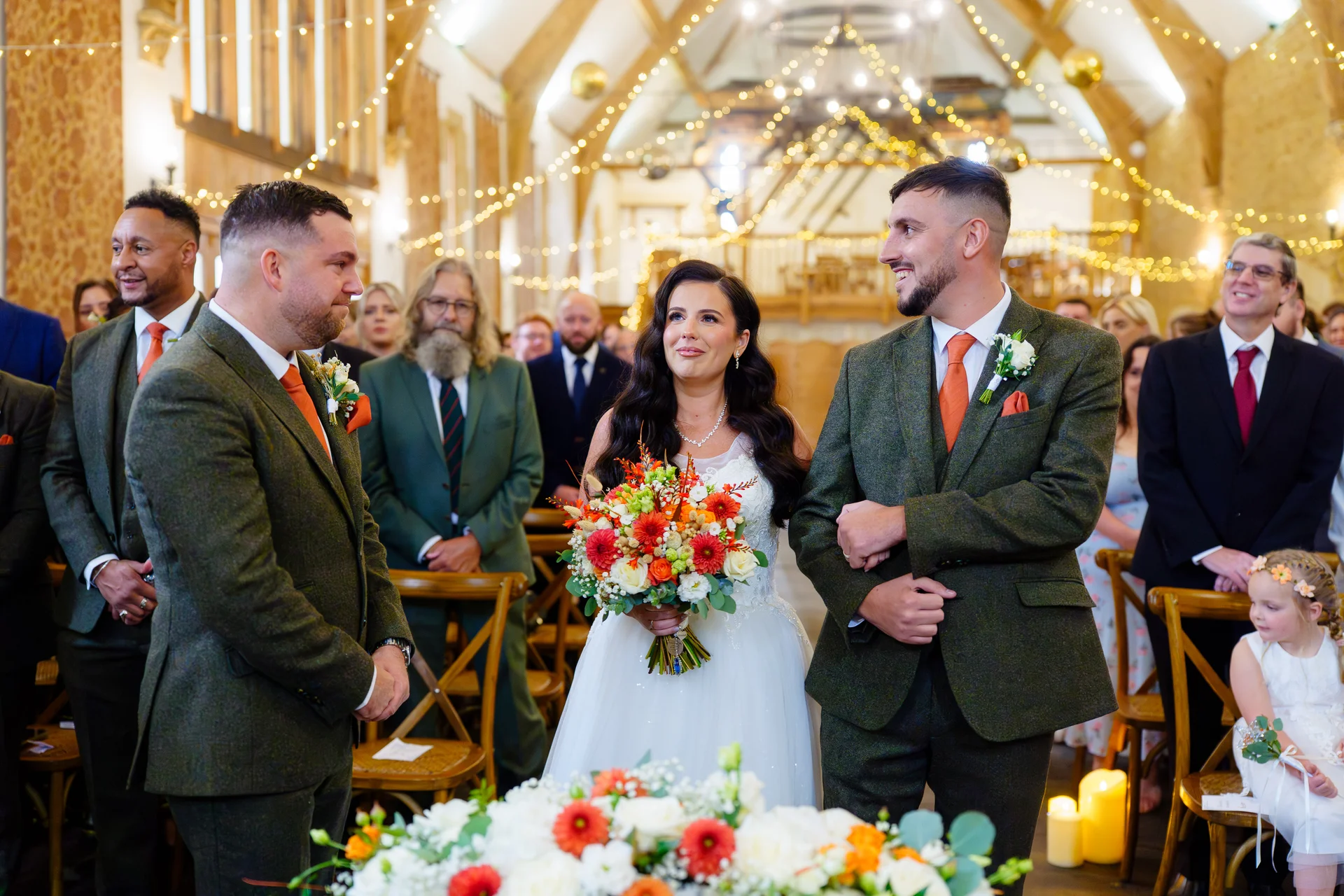 A smiling bride holding an autumnal bouquet stands at the altar with the groom and best man in tweed suits inside a rustic barn decorated with fairy lights.