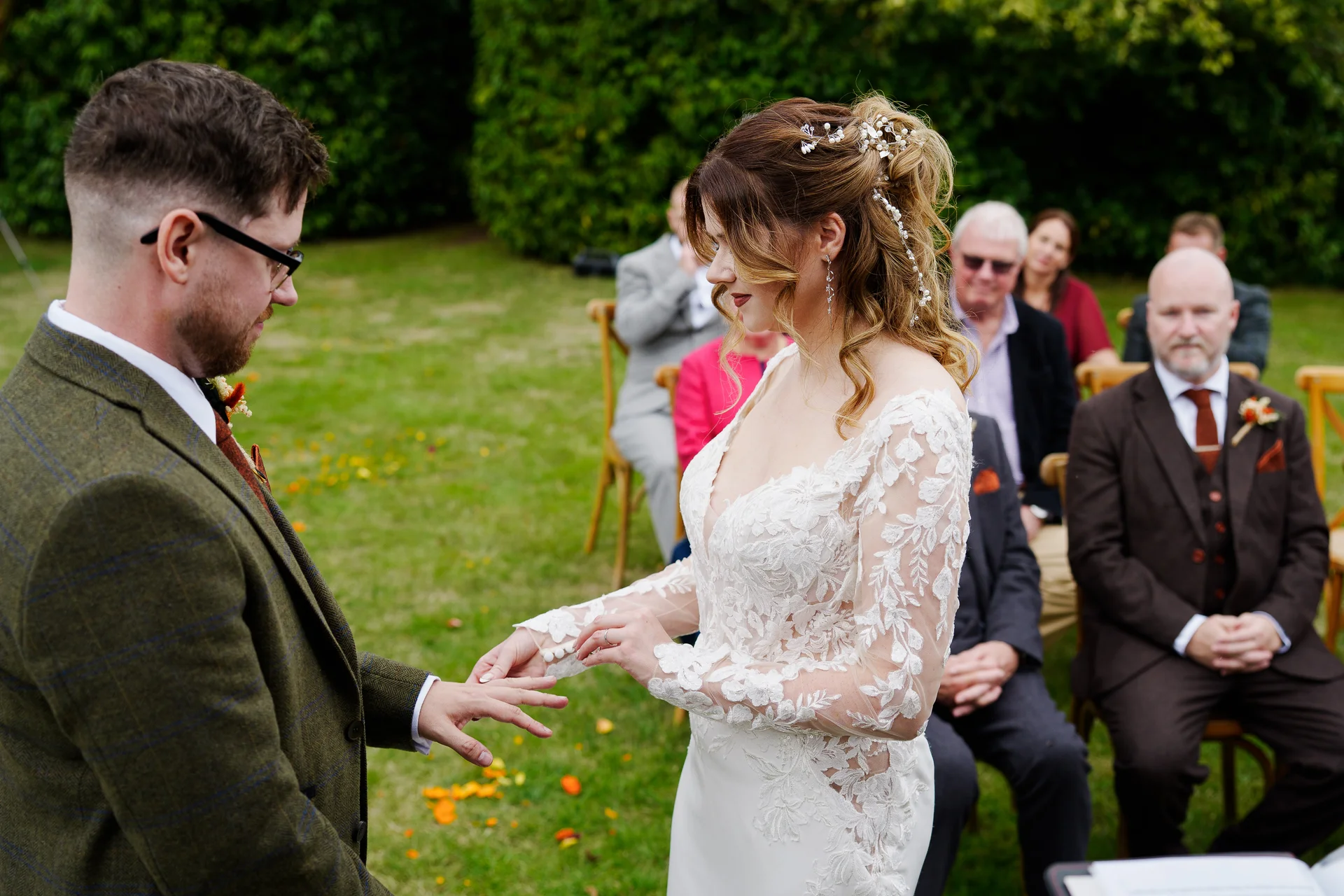 The groom gently places a wedding ring on the bride's finger during an outdoor garden ceremony, surrounded by seated guests.