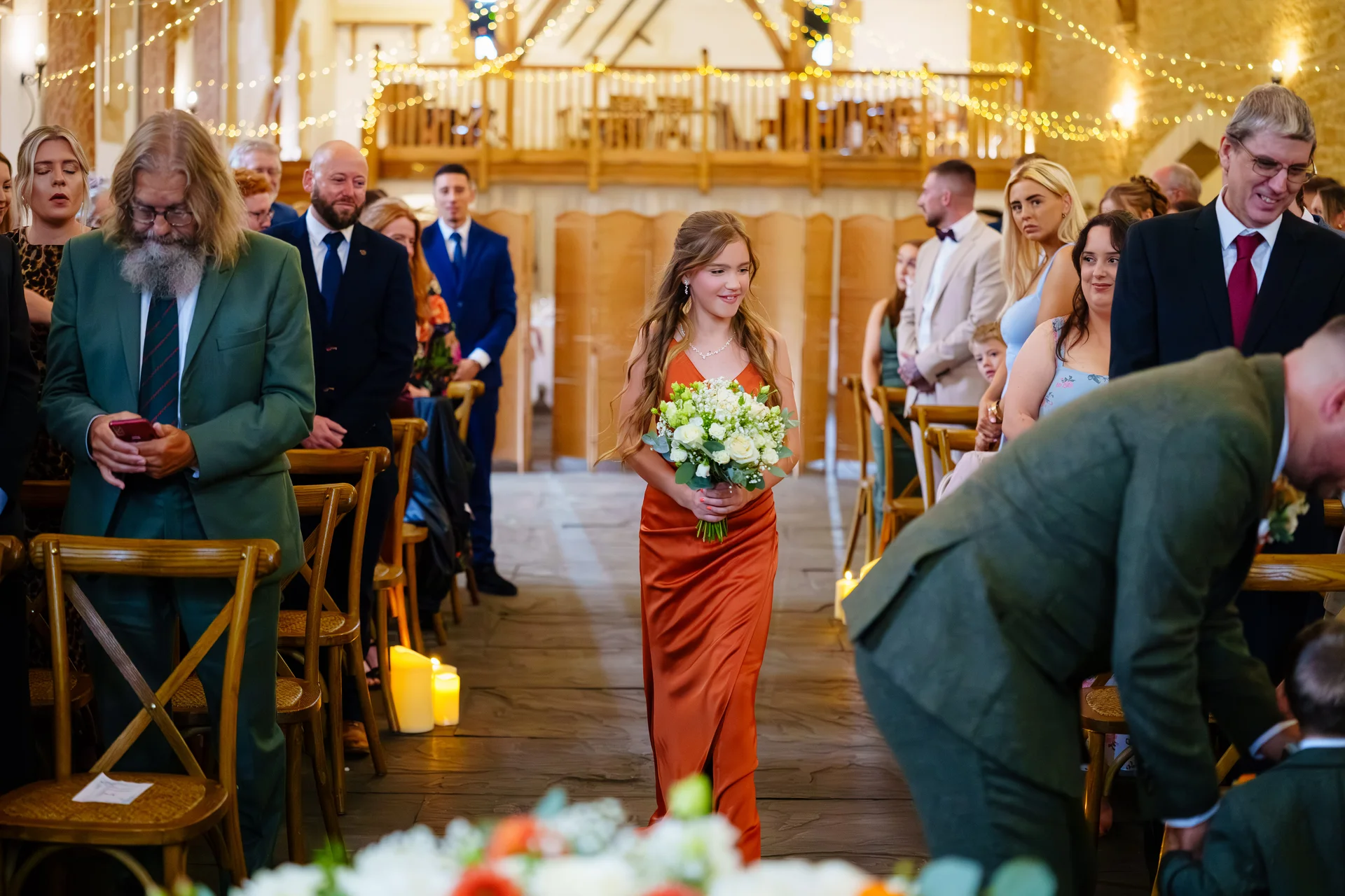 A bridesmaid in a rust-orange dress holding a bouquet walks down the wooden aisle of a rustic barn venue decorated with fairy lights and candles.