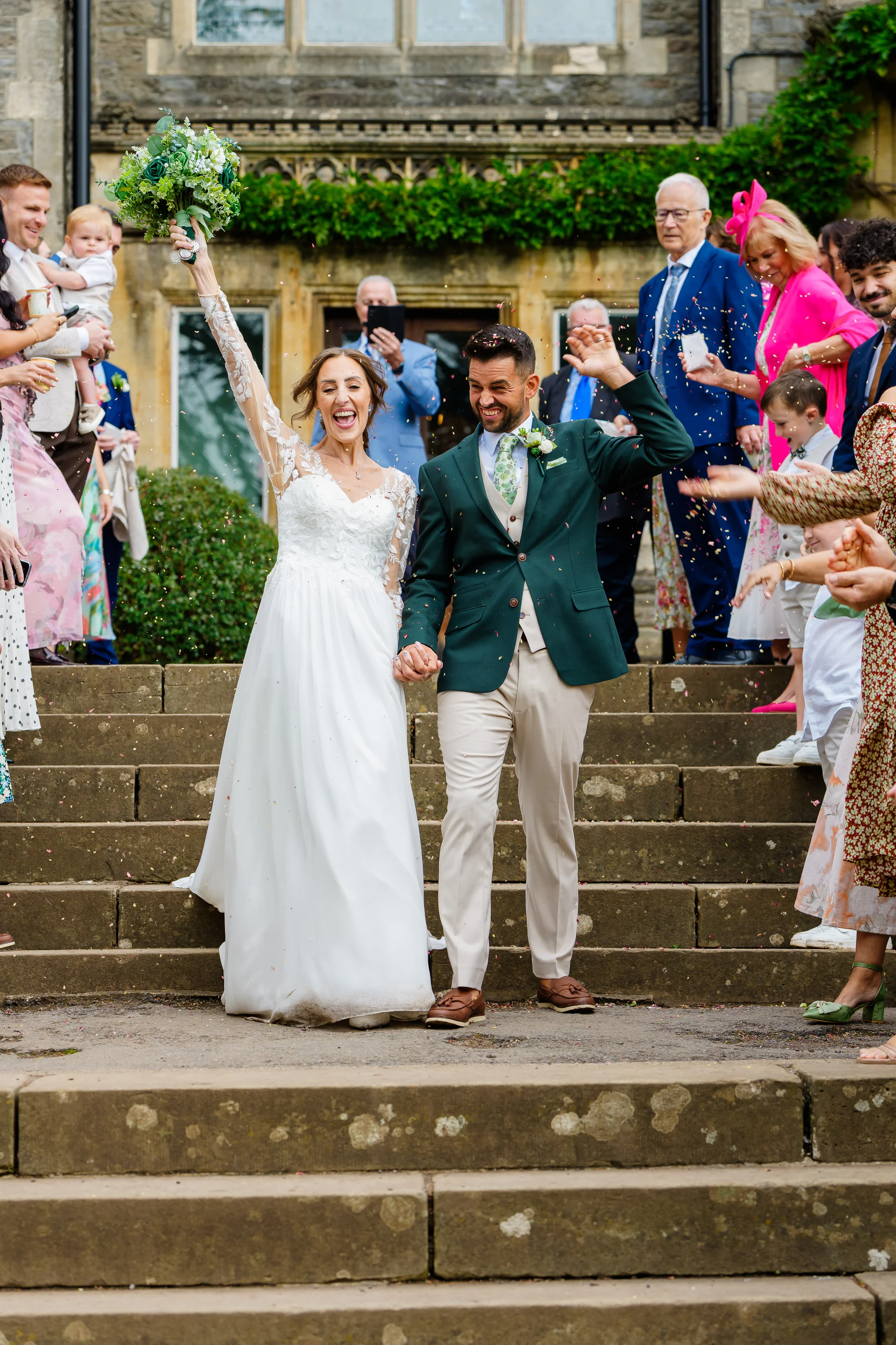 Newlyweds walk down stone steps holding hands while guests shower them with colorful confetti outside a historic country house venue.