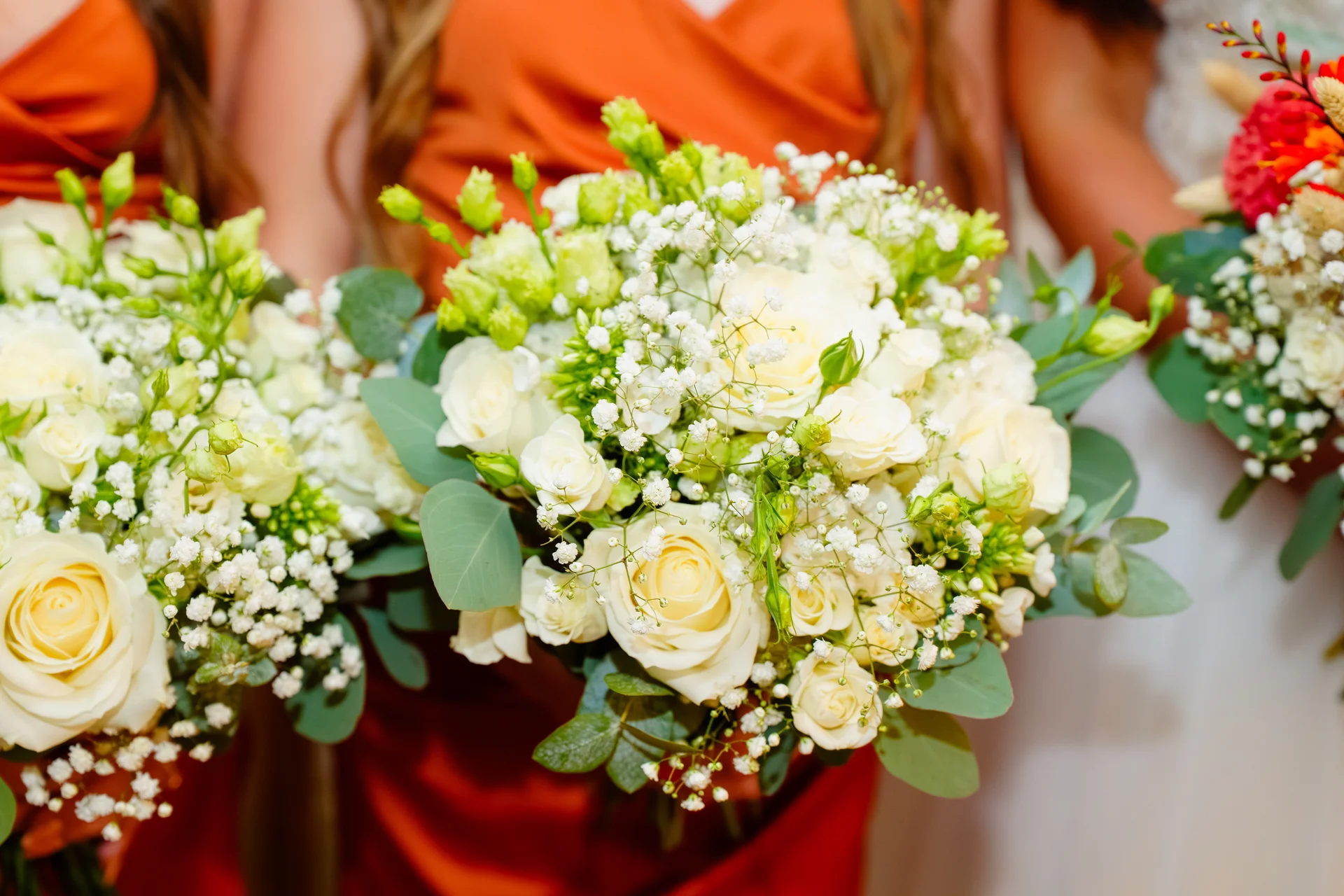 Bridesmaids holding lush hand-tied bouquets featuring white roses, delicate baby's breath, and eucalyptus foliage against vibrant orange dresses.