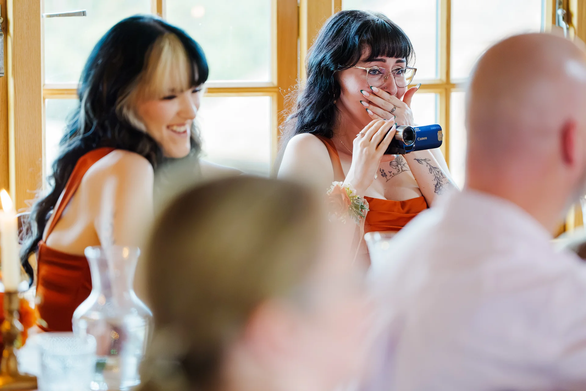 An emotional wedding guest holds her hand over her mouth while filming with a vintage camcorder during the reception speeches.