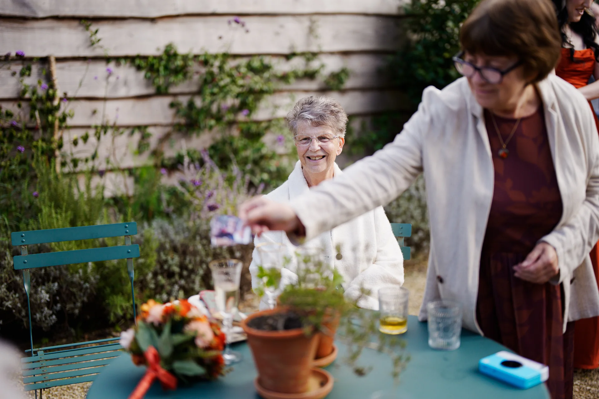 An older female guest smiles warmly at an outdoor wedding table setting while another guest reaches across with a disposable camera in a natural, candid moment.