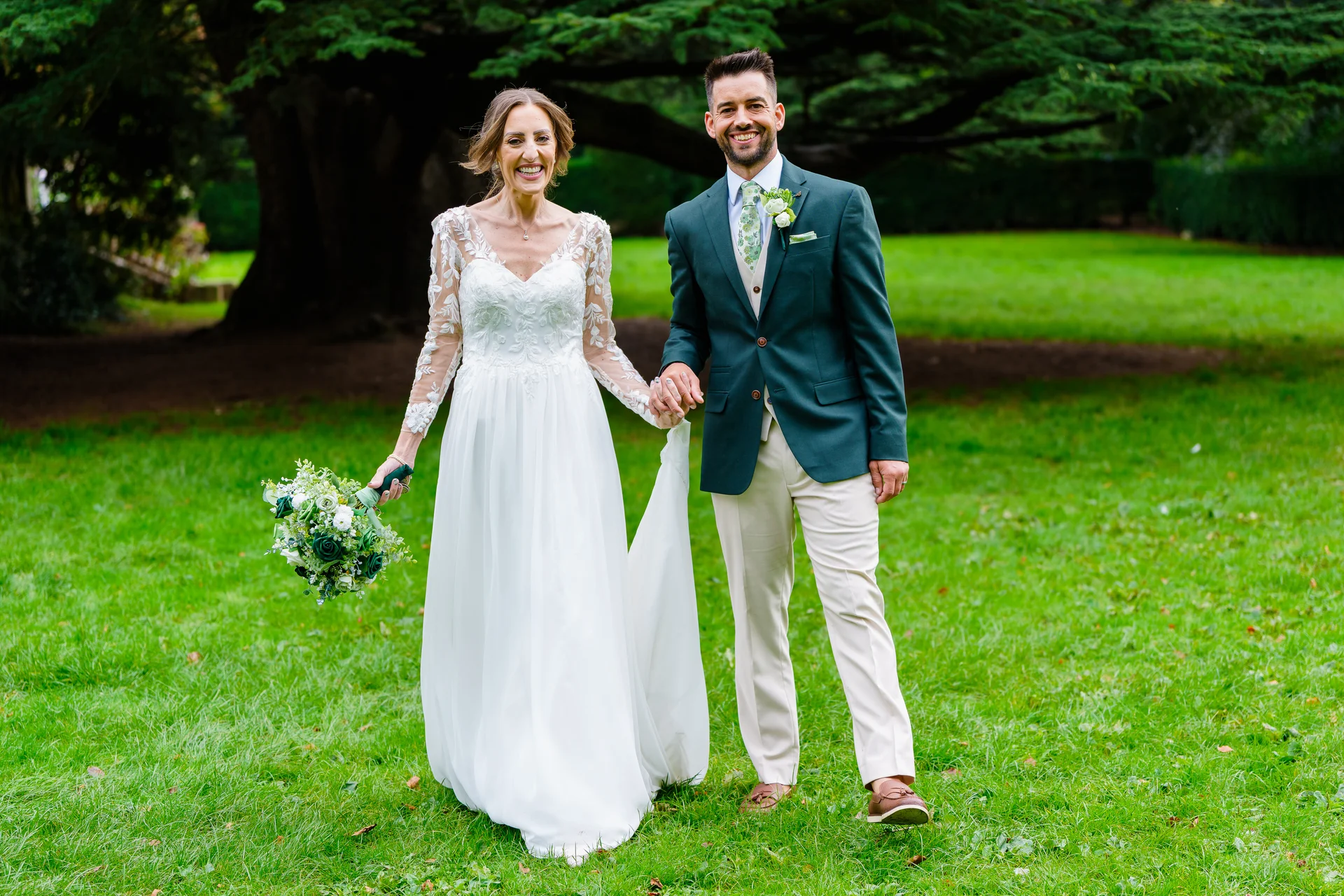 A joyful newlywed couple walks hand-in-hand across a lush green lawn in front of a large tree, with the bride holding a bouquet and wearing a lace-sleeved gown.