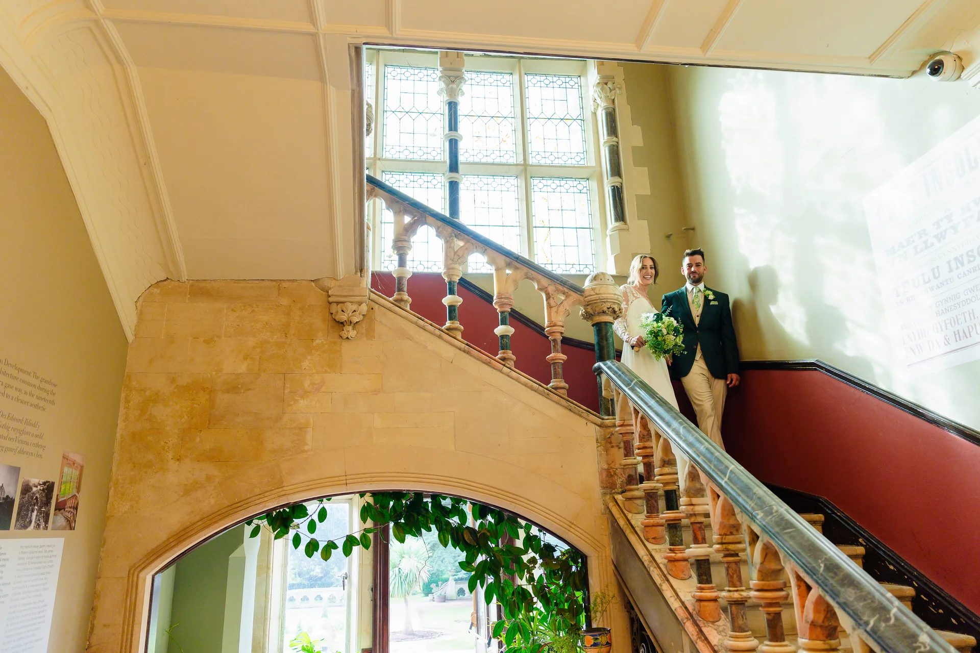 A newlywed couple poses on a grand stone staircase featuring an ornate balustrade and large stained glass windows in a historic manor house venue.