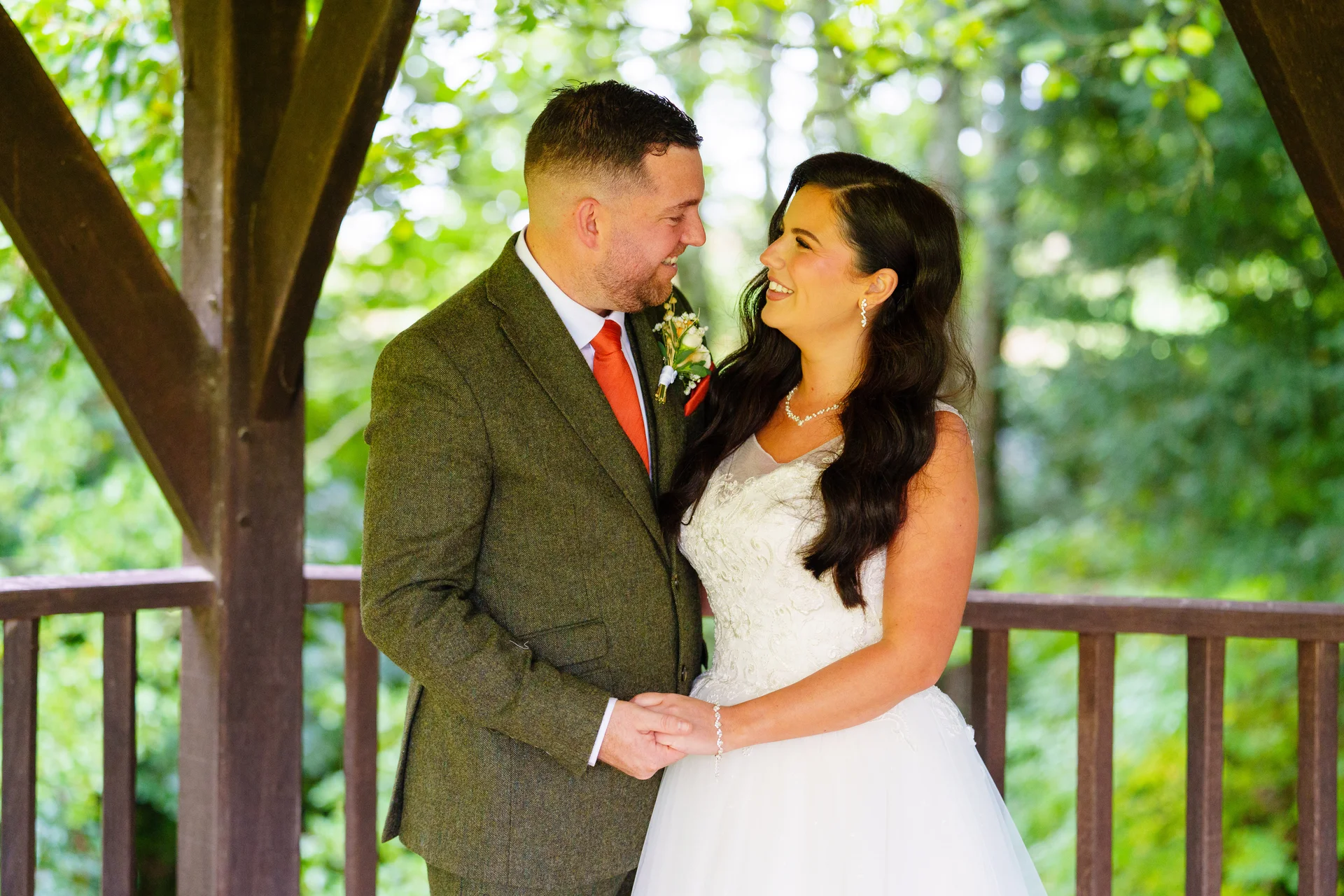 A smiling bride and groom hold hands while standing inside a wooden gazebo surrounded by lush green foliage on their wedding day.