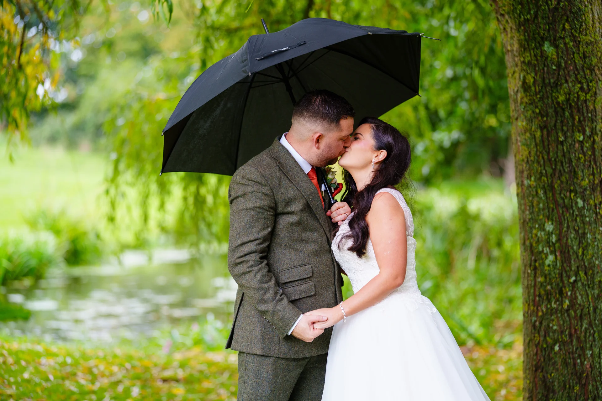 A bride and groom share a romantic kiss under a black umbrella by a tranquil lakeside with lush green trees.