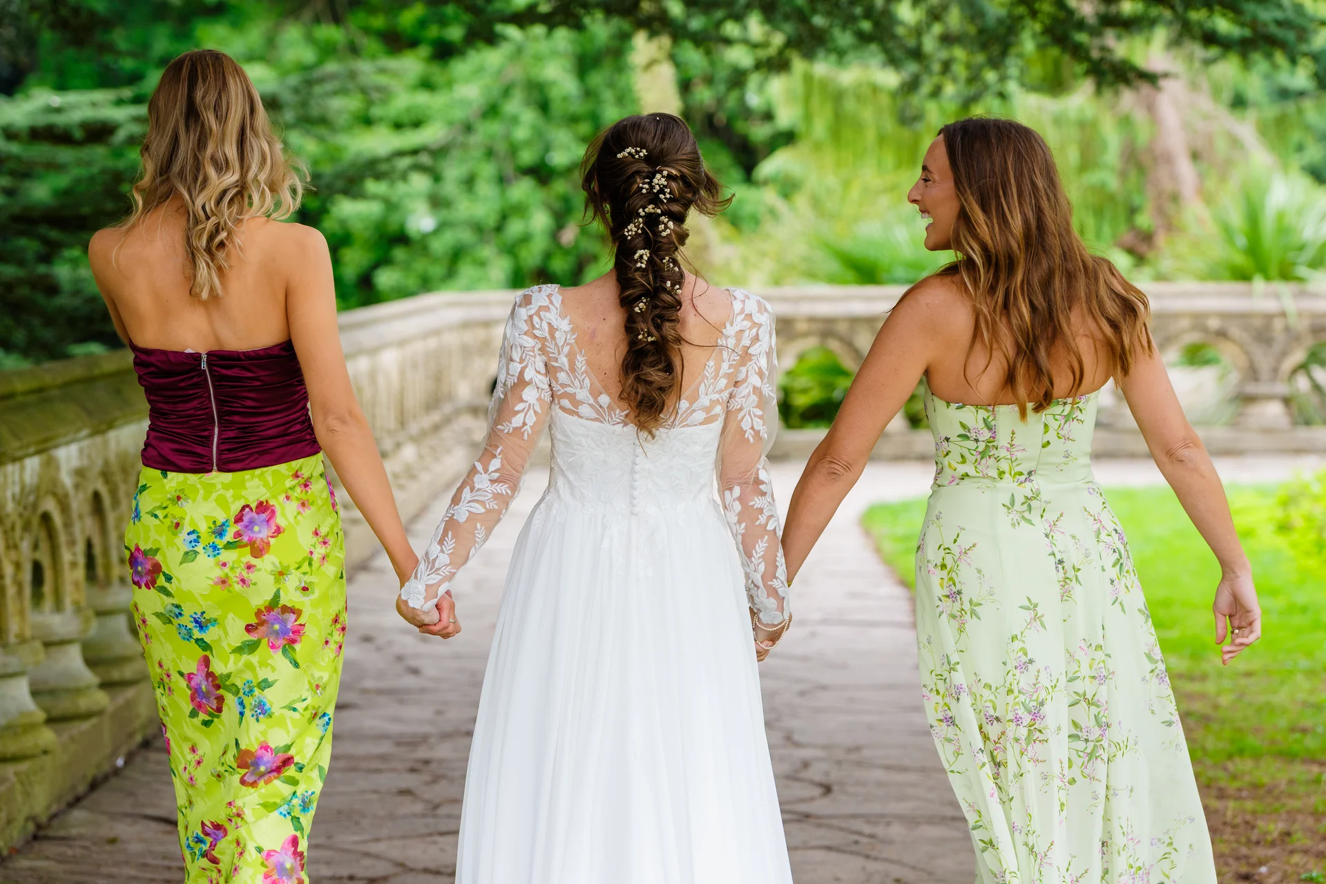 A bride in a long-sleeved lace gown walks hand in hand with two bridesmaids wearing mismatched floral dresses along an outdoor pathway.