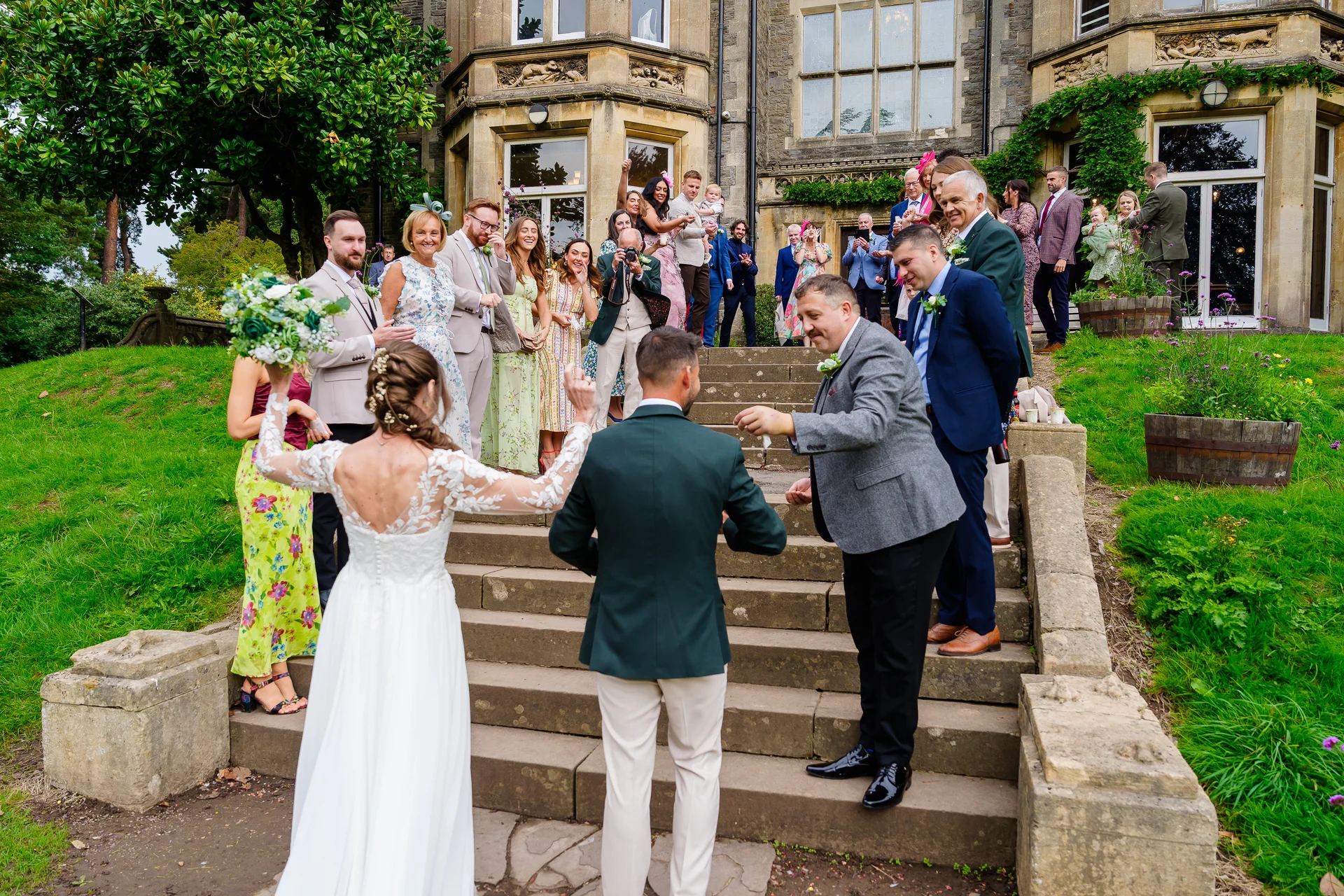 Newlyweds stand at the bottom of grand stone steps outside a country house, facing family and friends who cheer and throw confetti from above.