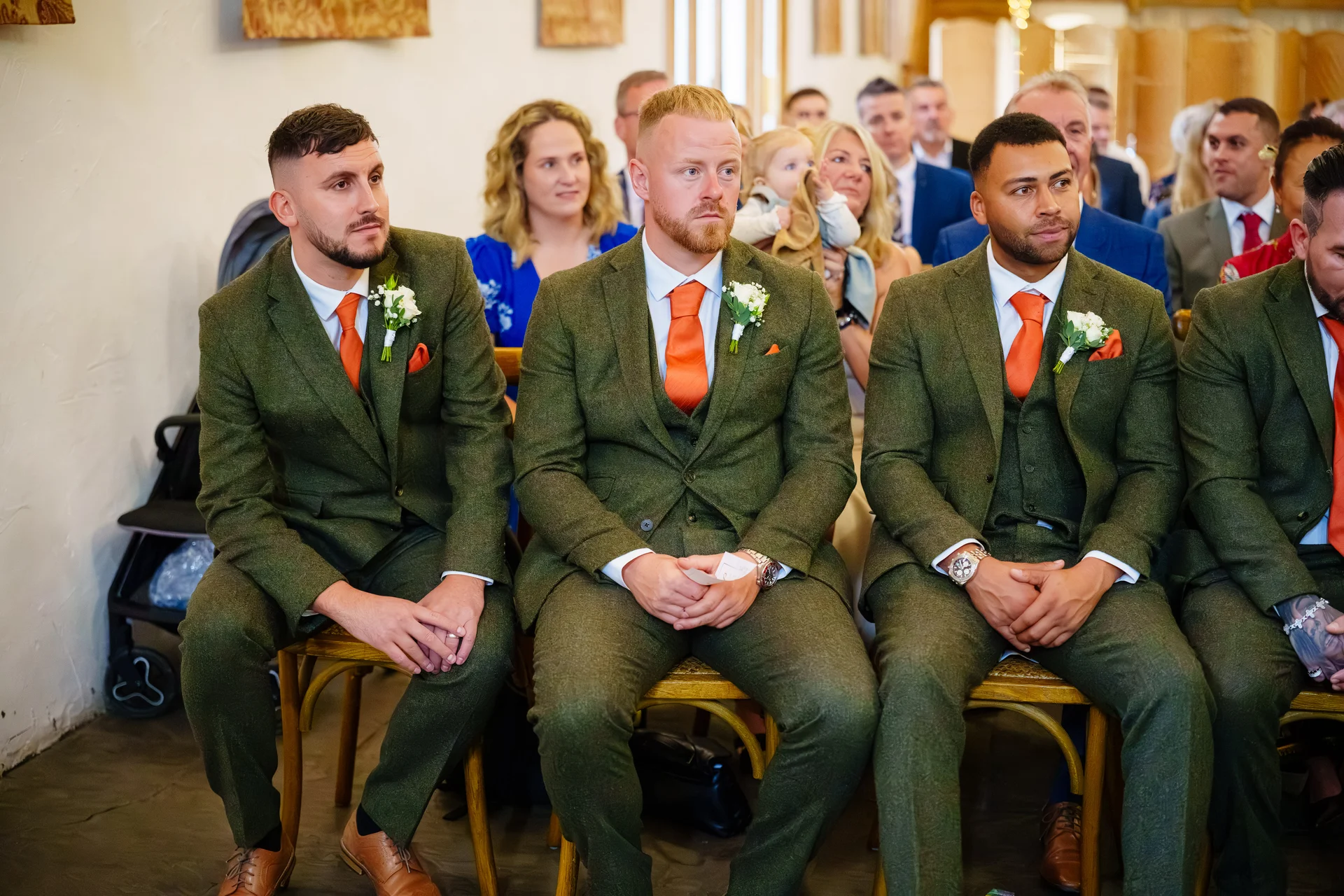A group of groomsmen wearing matching green tweed three-piece suits with vibrant orange ties and pocket squares seated together waiting for the ceremony.