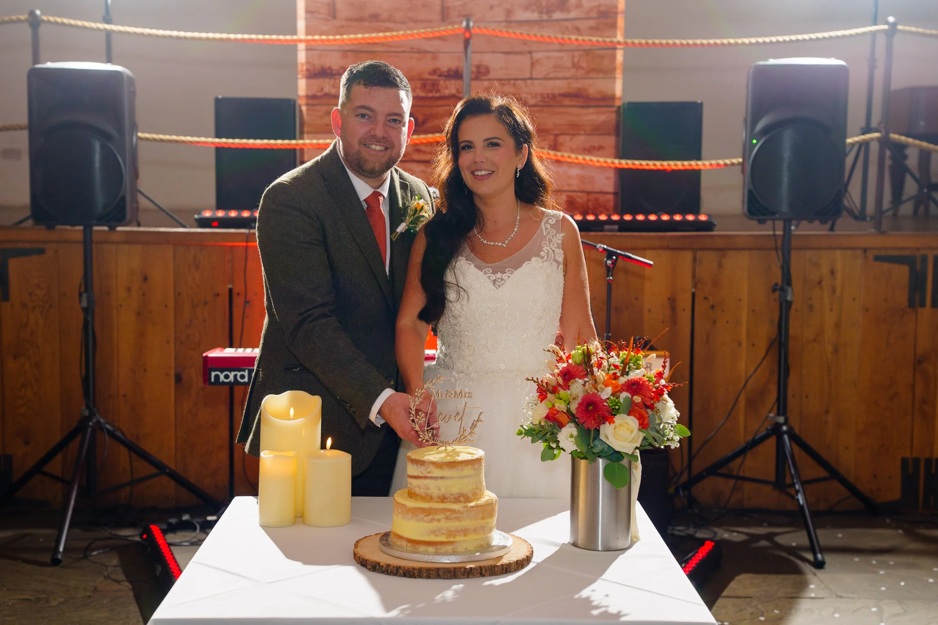 A smiling bride and groom in a rustic barn venue proudly cut their semi-naked wedding cake with a custom wooden topper, set against a stage with musical equipment and lighting.