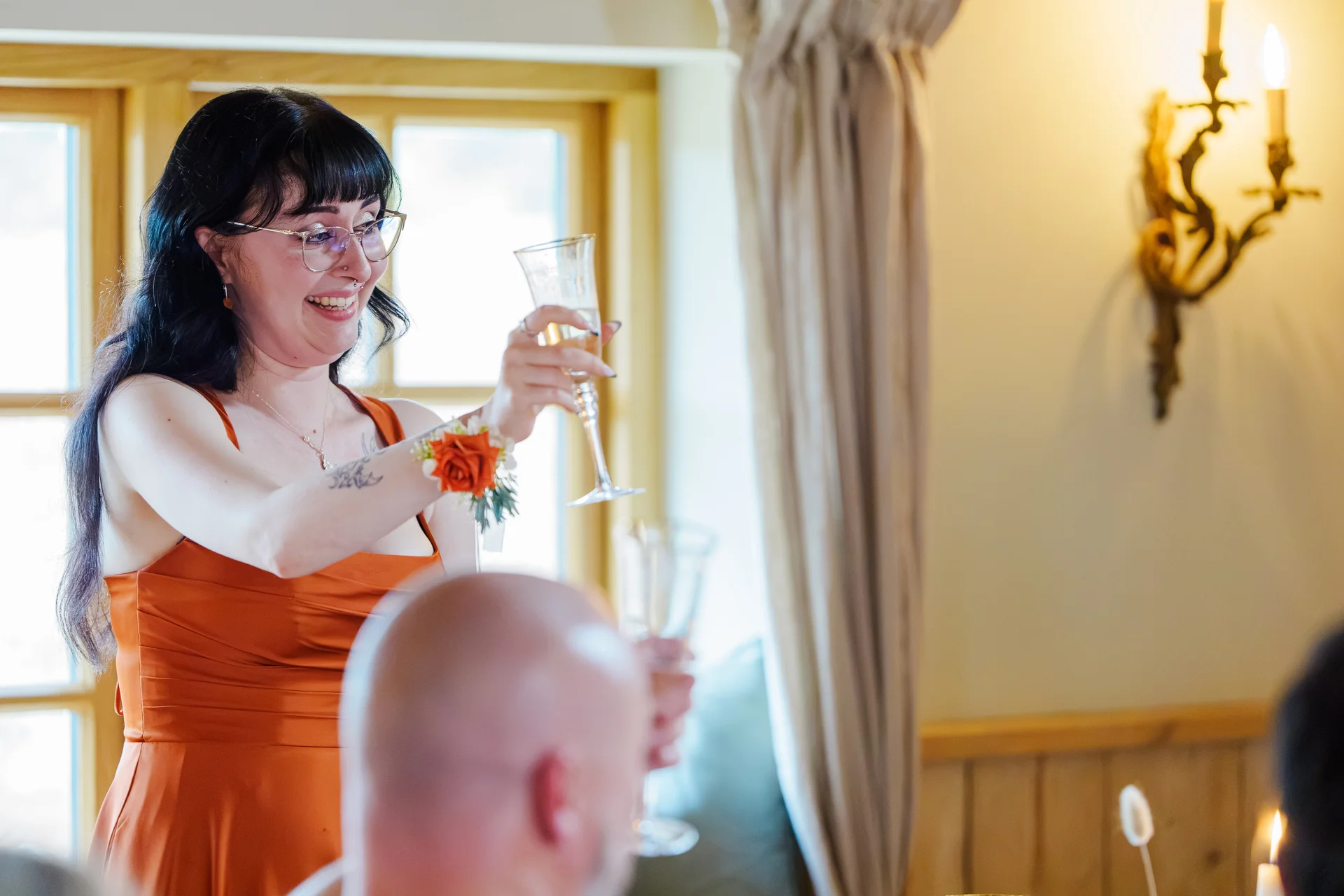 A smiling woman wearing glasses and an orange dress raises a champagne flute during the wedding speeches.