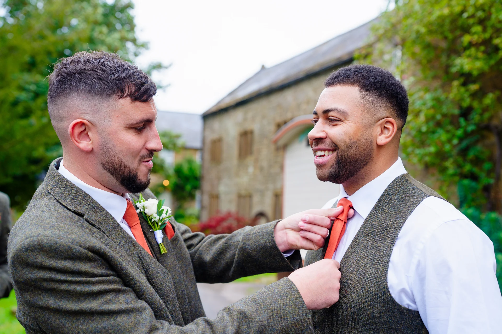 Two grooms in tweed waistcoats and burnt orange ties share a joyful moment outdoors outside a stone barn venue.