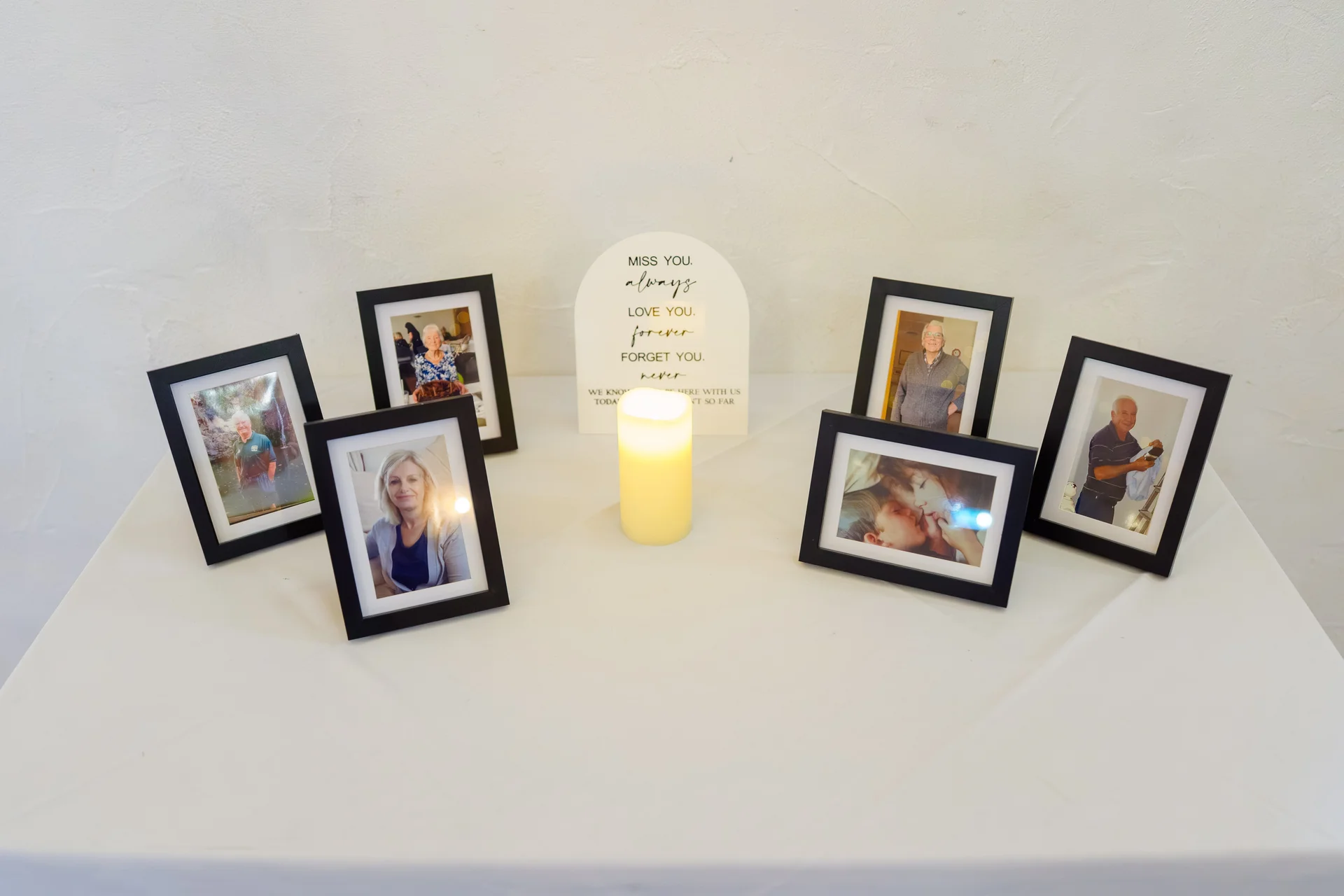 A thoughtful memory table features several framed family photographs arranged around a glowing memorial candle and a commemorative sign on a white tablecloth.