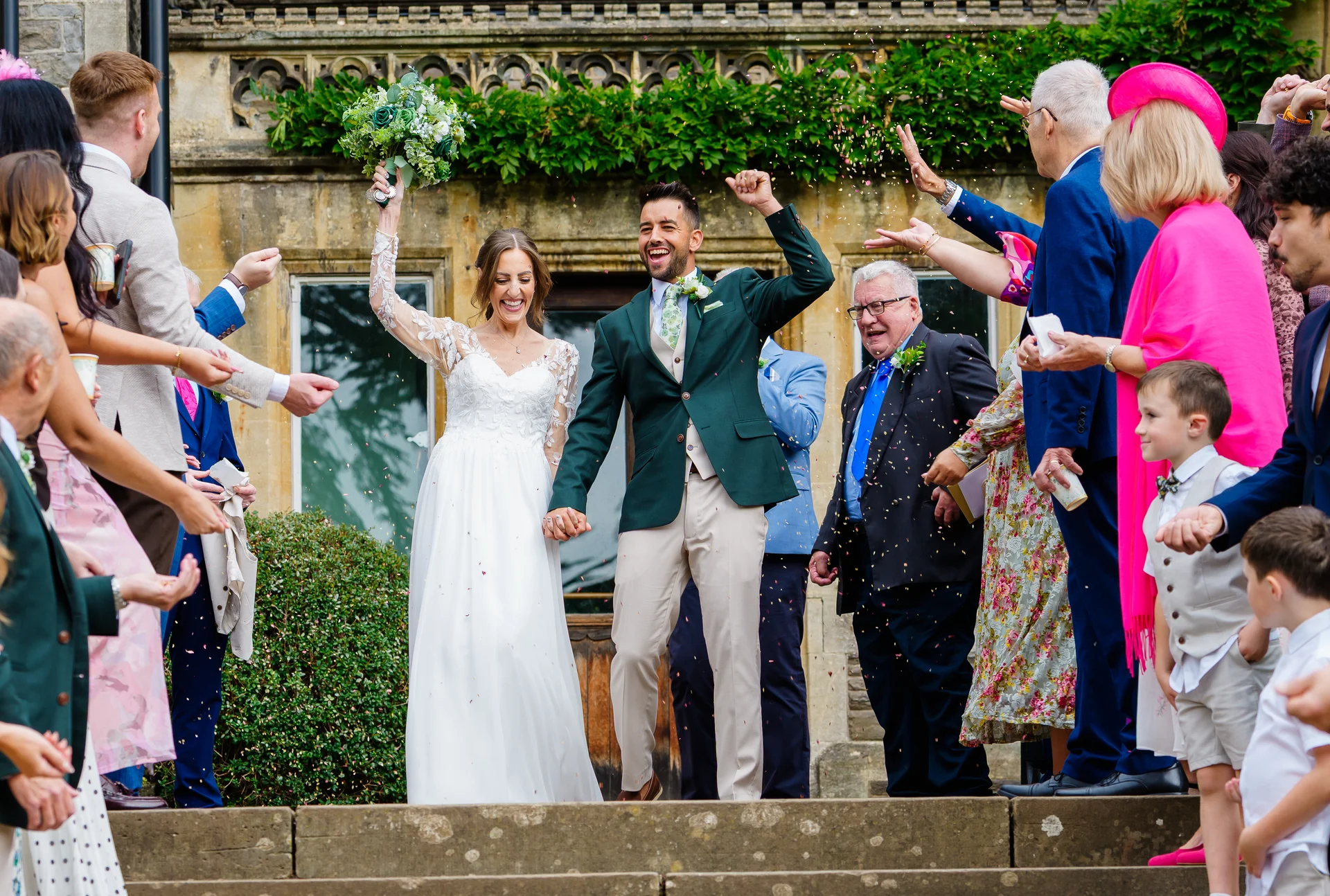 A joyful newlywed couple walks hand in hand down stone steps while celebrating guests shower them with colourful confetti outside a stone country house.