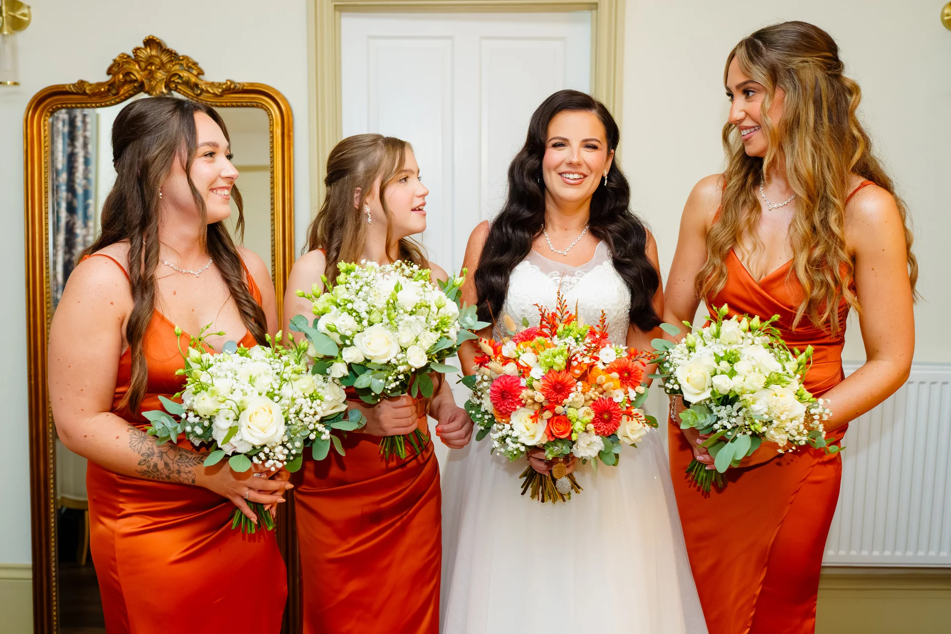 A smiling bride stands holding an autumnal floral bouquet alongside three bridesmaids dressed in matching rust satin gowns and holding white and green bouquets.