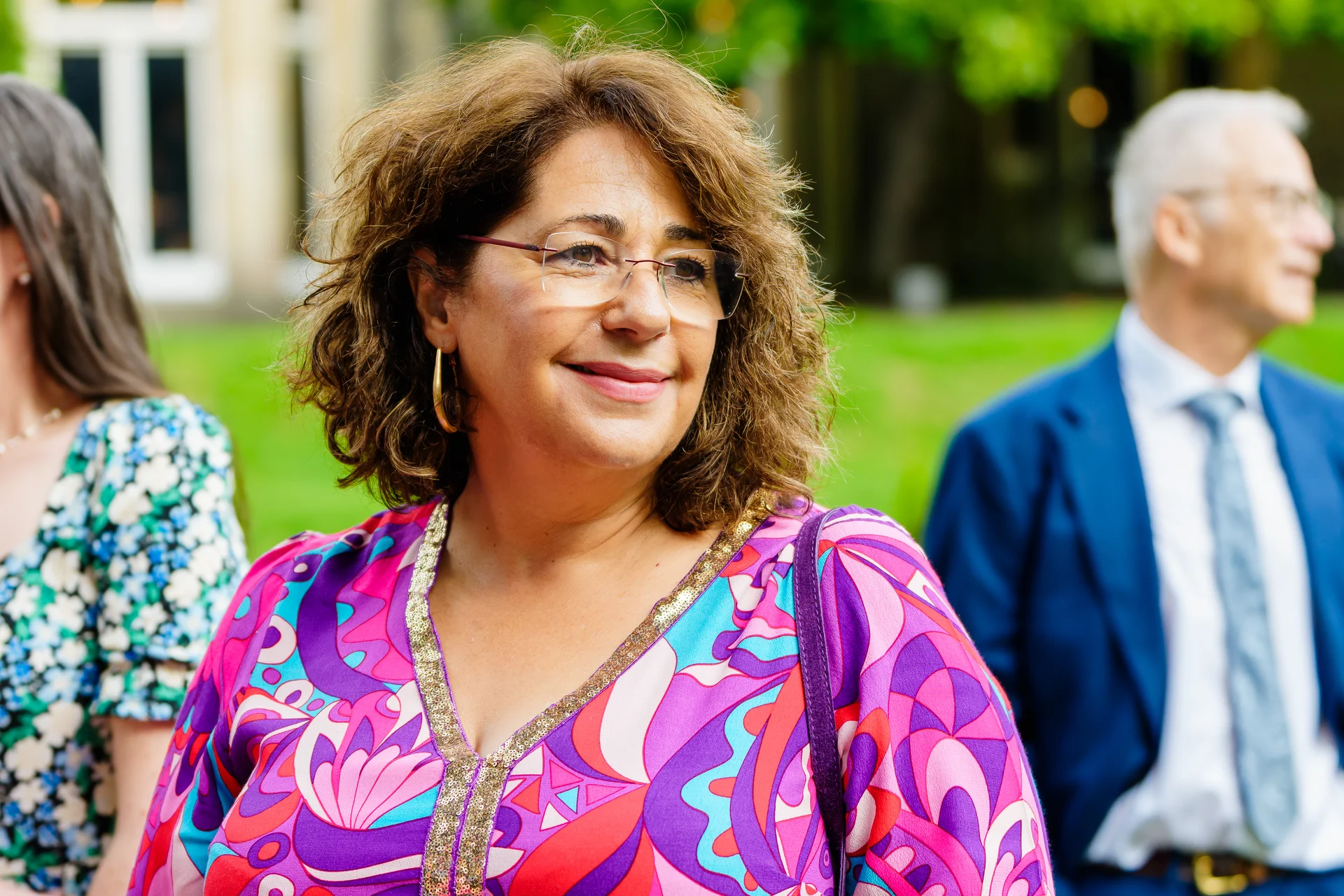 A wedding guest with curly brown hair wearing a colourful patterned dress smiles outdoors during the celebration.