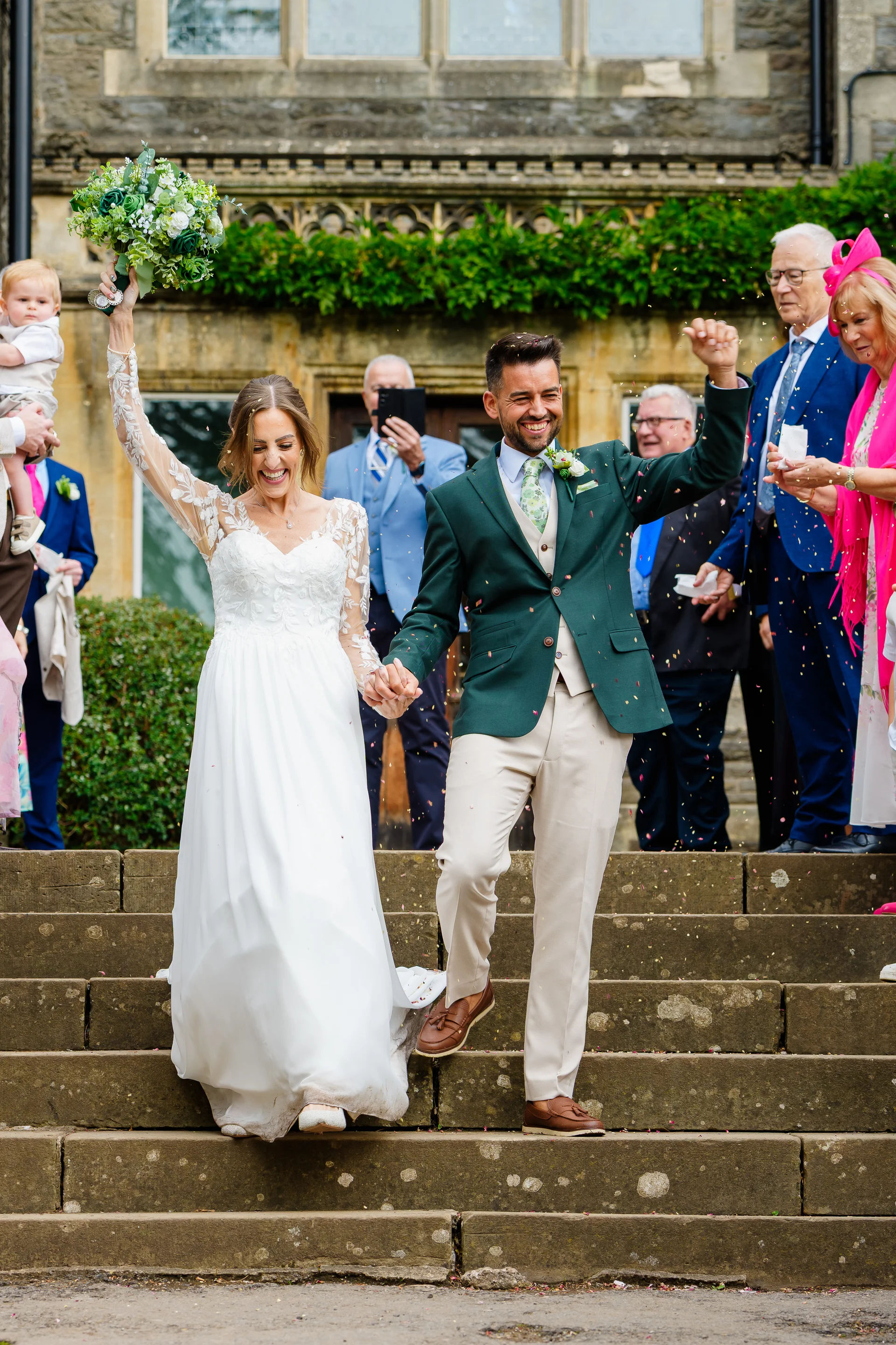 A newlywed couple joyfully walk down stone steps outside a country house venue while guests shower them with colorful confetti.